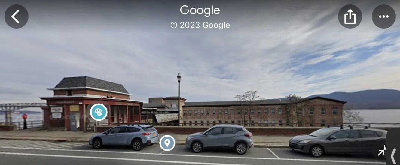Street view showing a historical building, parked cars, and a waterfront in the background with mountains and a cloudy sky.