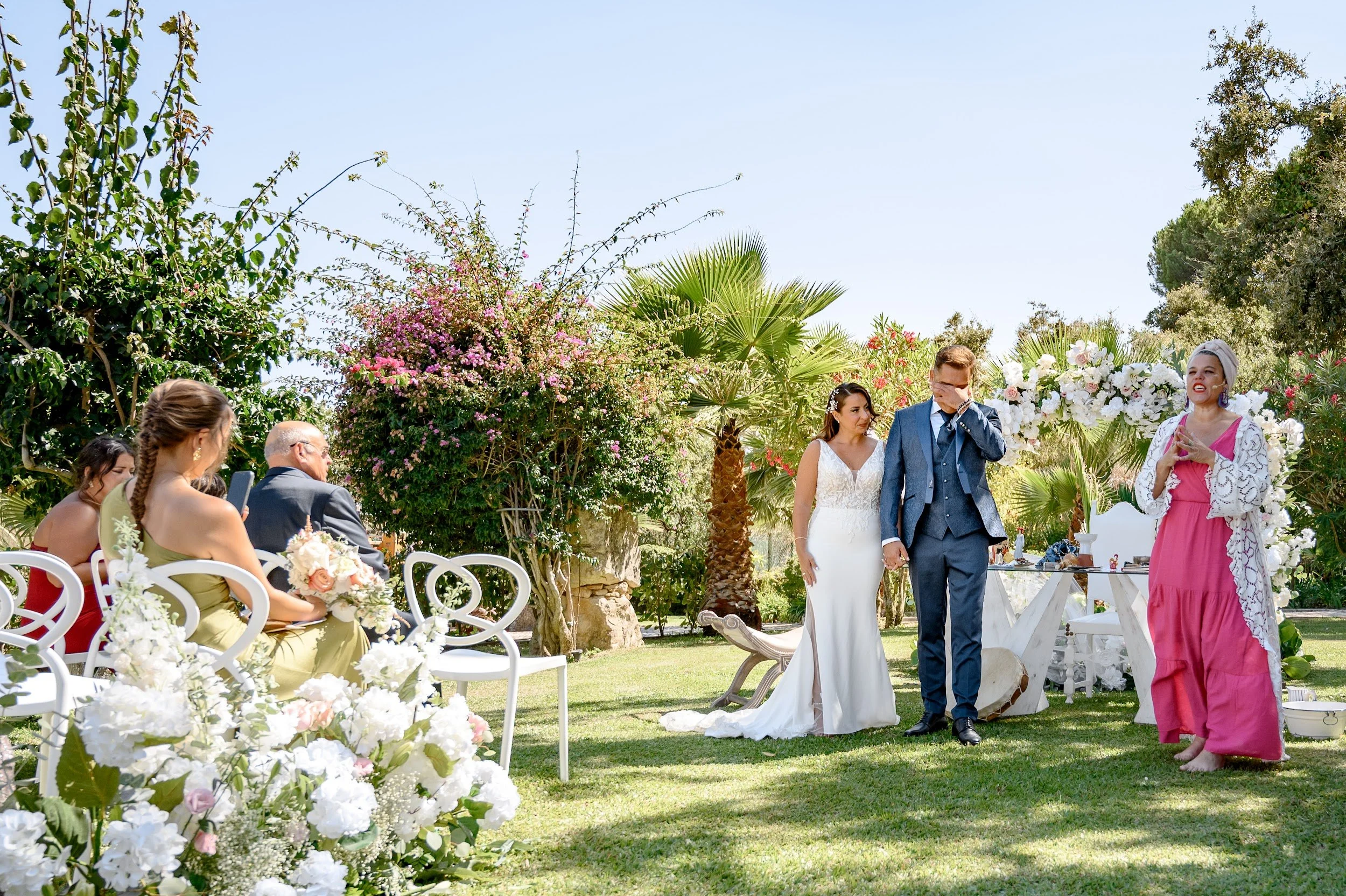 Casamento ao ar livre com noivos no centro, ao lado de uma mulher em vestido rosa, rodeados de convidados sentados, com cortinas brancas e muitas flores brancas e rosas, árvores e céu azul ao fundo.