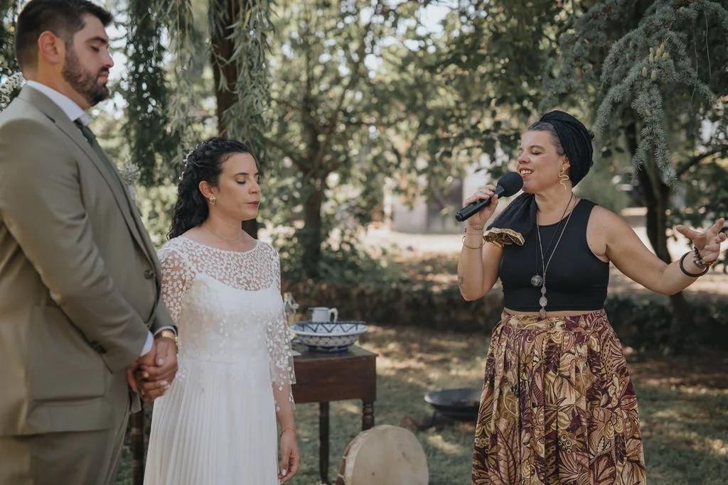 Três pessoas em um casamento ao ar livre, onde um casal está de frente para uma mulher que está cantando ou falando ao microfone.