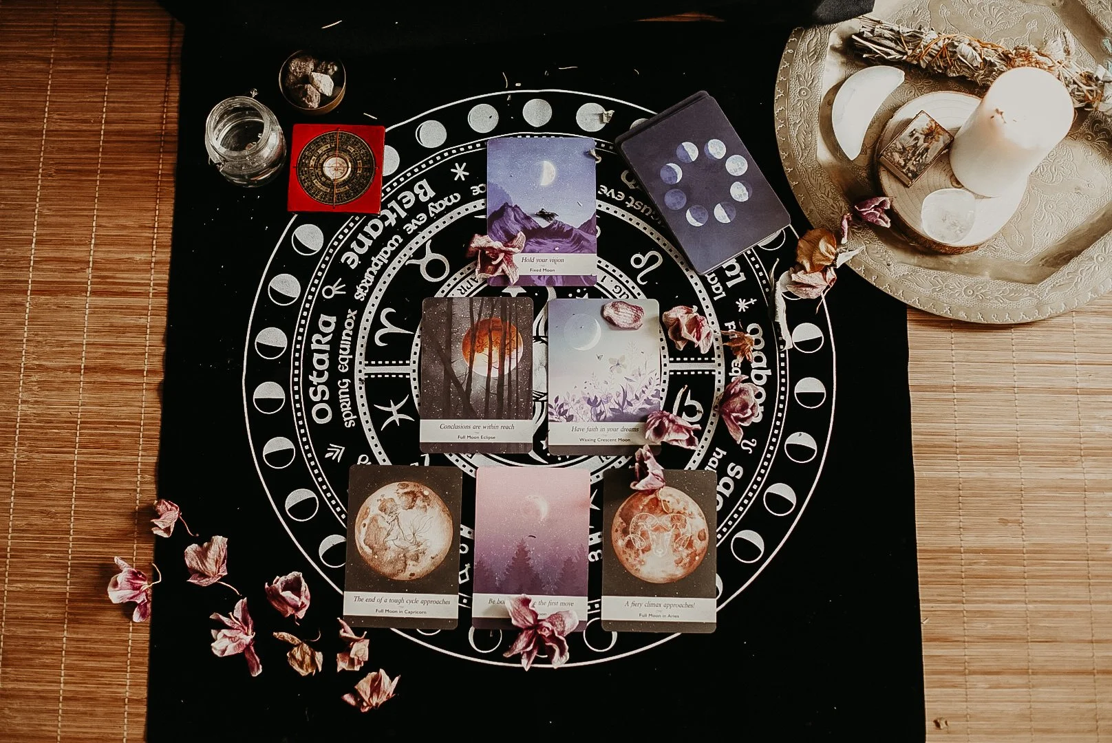 Tarô de cartas sobre um tapete preto com símbolos astrológicos e fases da lua, decorado com pétalas secas, ao lado de uma vela, cristal e outros objetos ritualísticos.