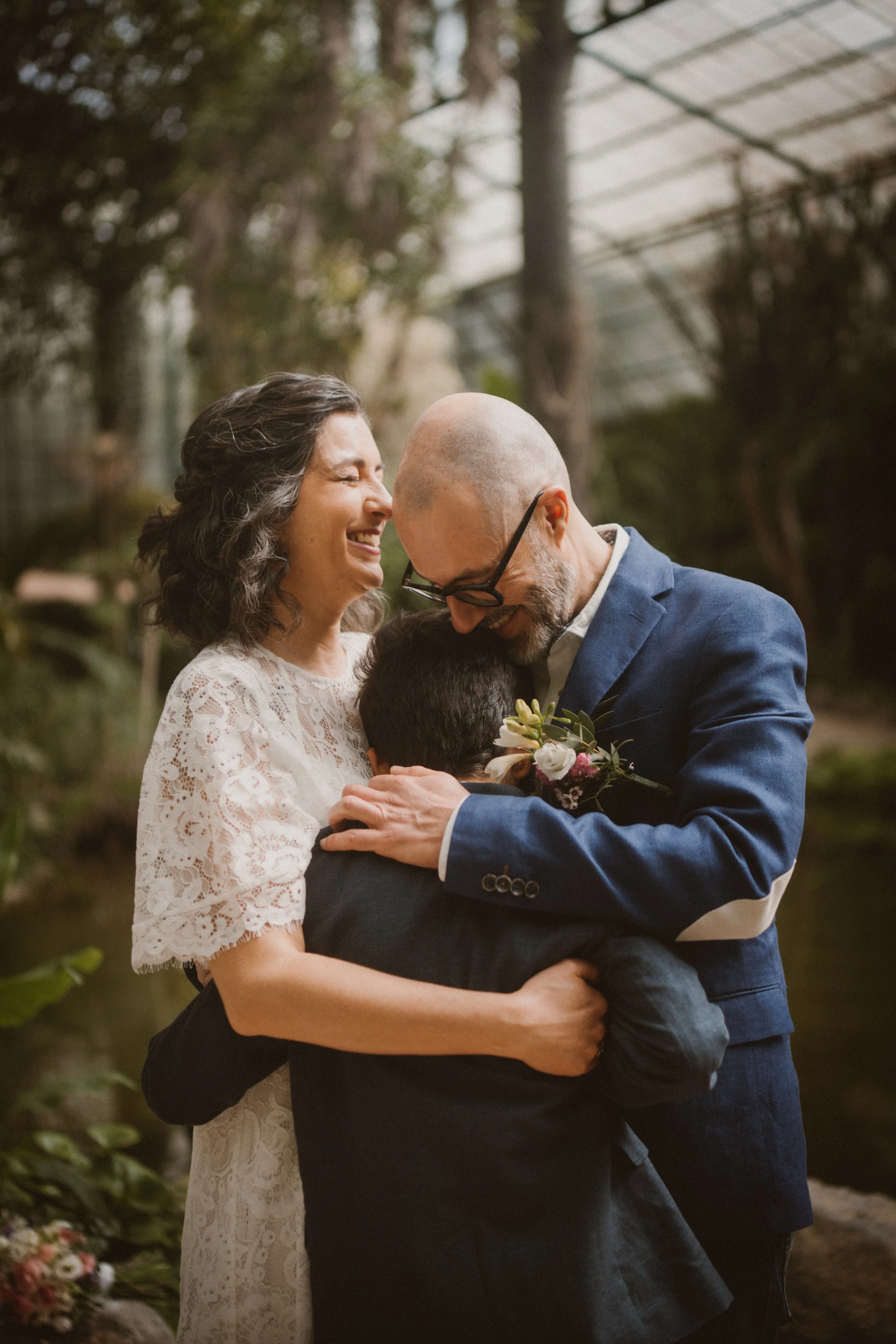 Casal sorridente em casamento, abraçando uma criança, ao ar livre com árvores ao fundo.