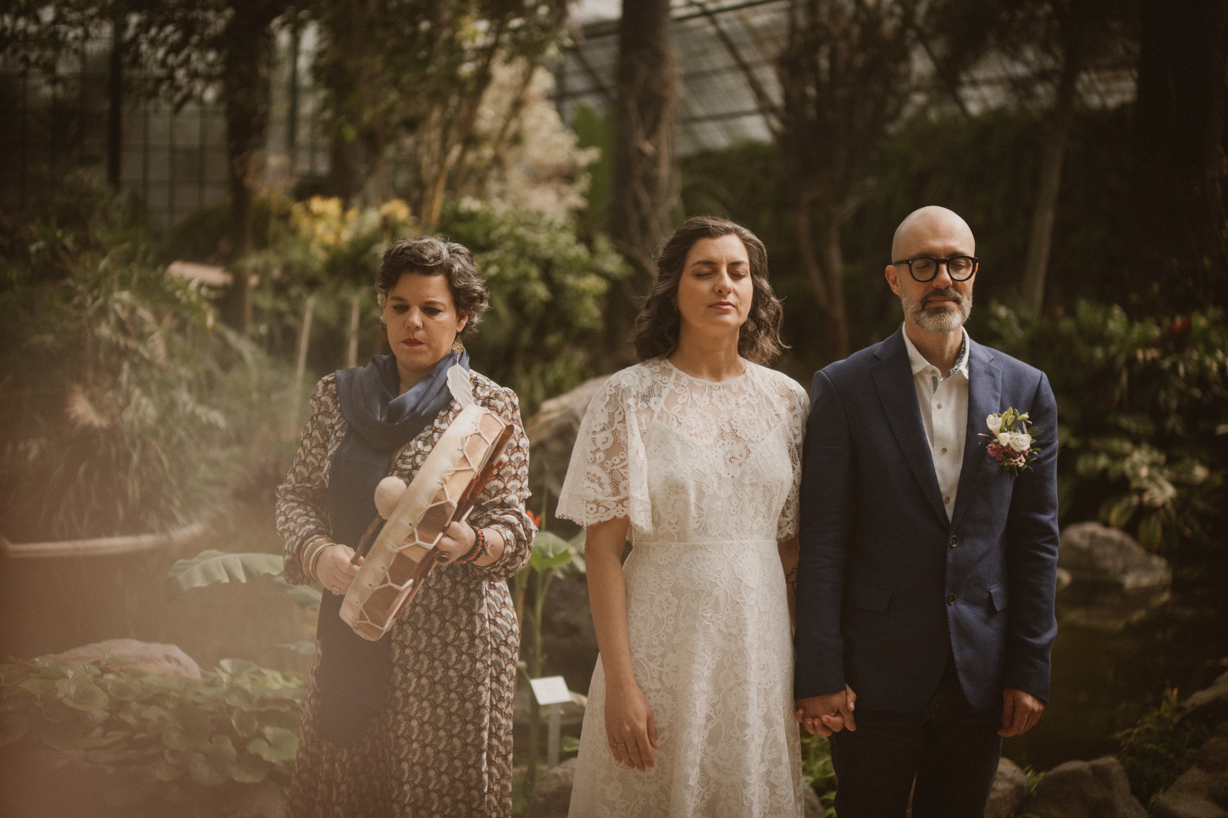Três pessoas, uma mulher usando vestido de casamento, uma outra mulher e um homem com terno, segurando as mãos, participando de cerimônia ao ar livre em ambiente natural.