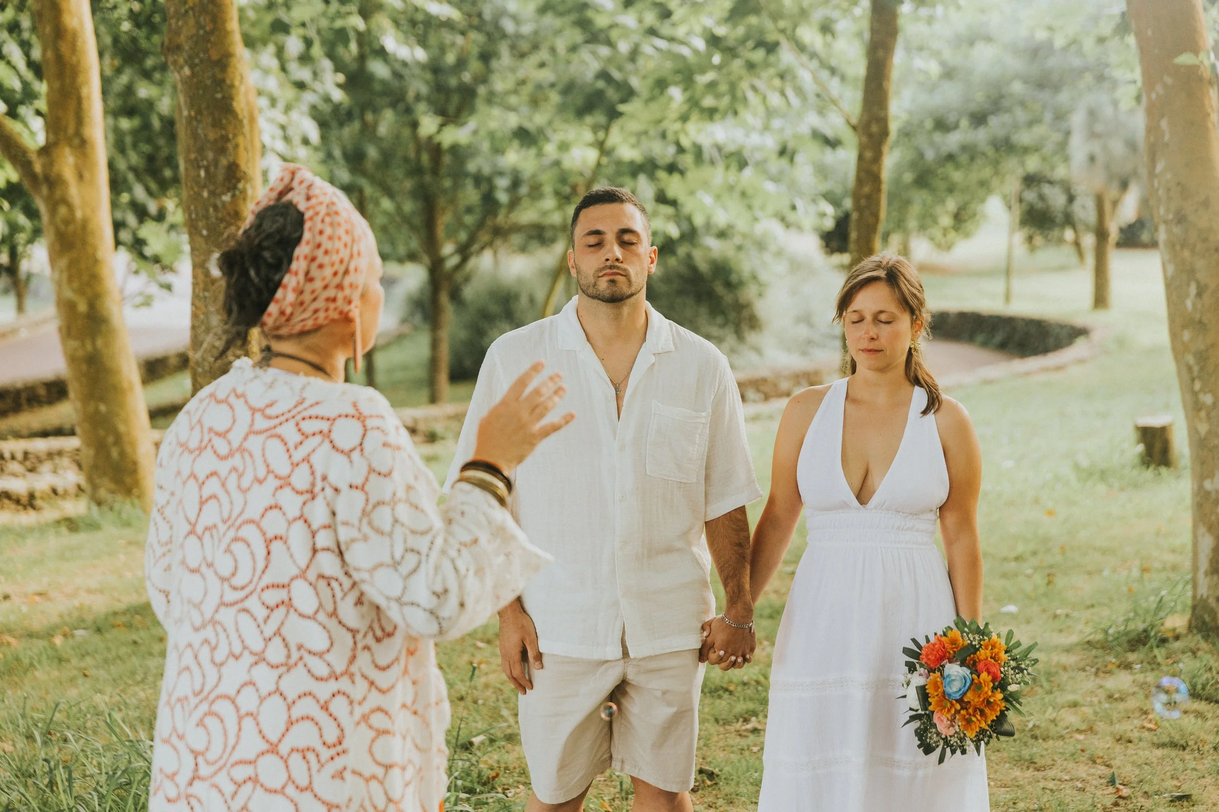 Casal fazendo cerimônia de casamento ao ar livre, com testemunha segurando as mãos do casal, que está de frente, com árvores ao fundo.