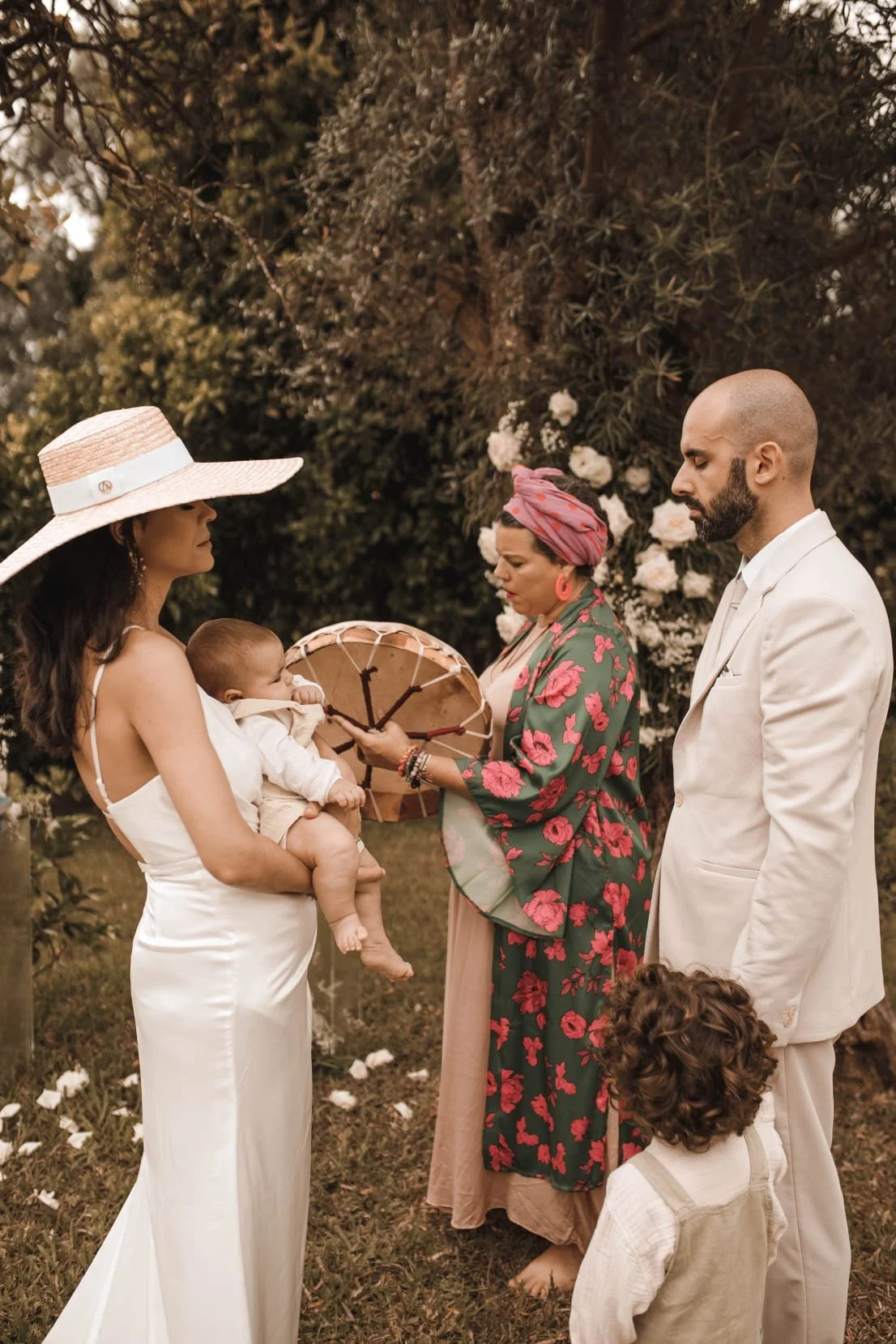 Casal na cerimônia de casamento ao ar livre, com a noiva segurando um bebê, cerca de duas pessoas de vestido tradicional, uma delas com um guarda-chuva, uma criança de costas ao lado do noivo, com flores ao fundo.