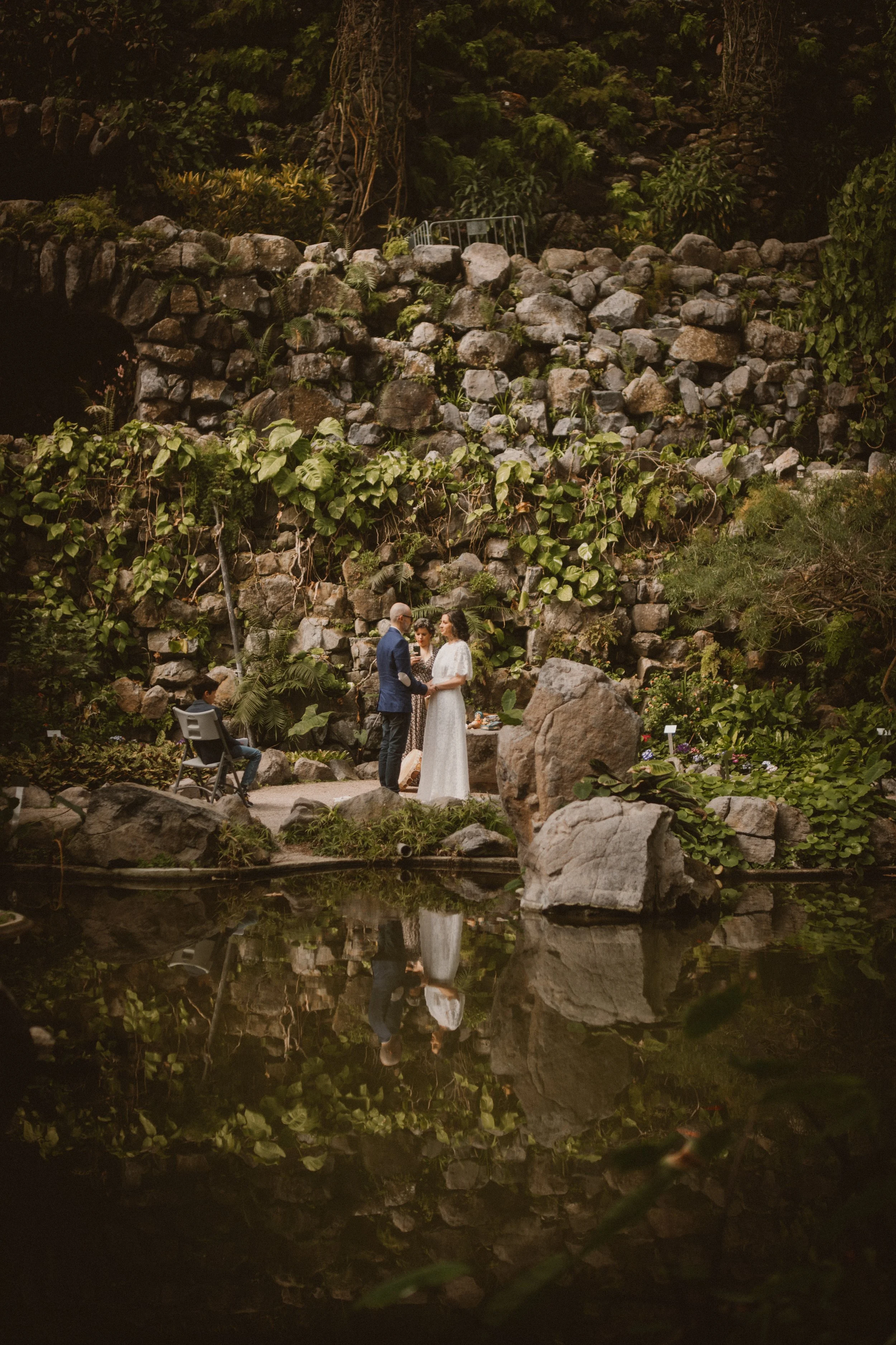 Casamento ao ar livre em um jardim ao entardecer, com um casal trocando votos em frente a um cenário de pedras e vegetação, refletido em um lago tranquilo.