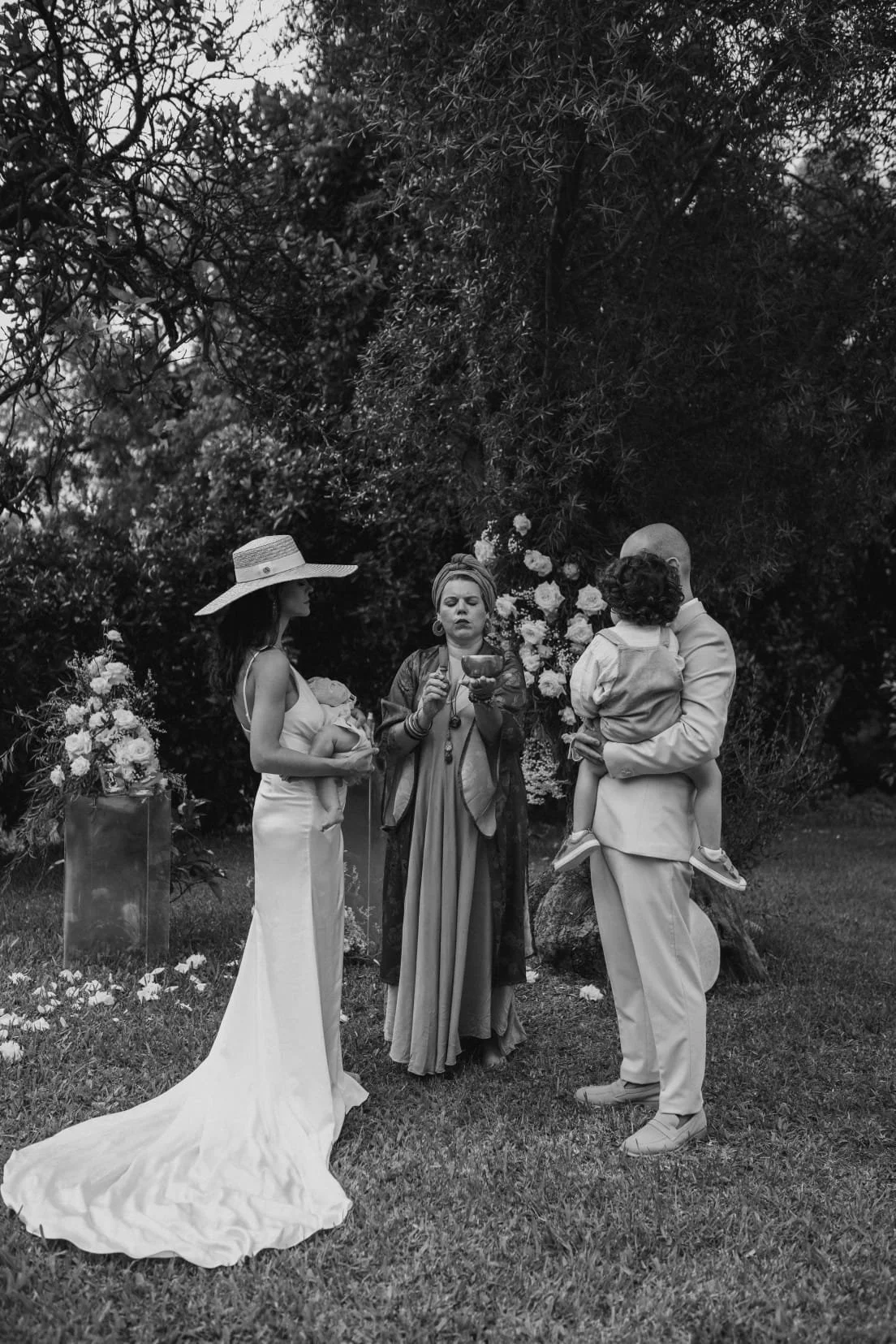 Cena de uma cerimônia de casamento ao ar livre com quatro pessoas, incluindo uma noiva, um homem e duas mulheres, uma delas segurando uma criança. Todos estão diante de um fundo de árvores e flores, em um ambiente natural.
