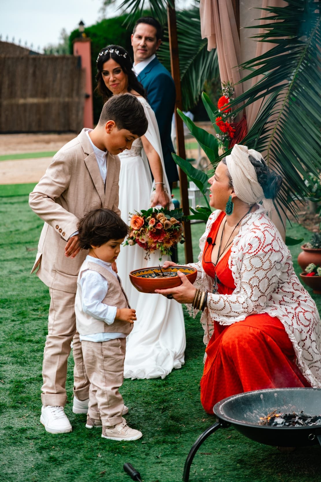 Pessoa vestida com roupas tradicionais indígenas entregando faca de depilação a uma criança na frente de outros adultos em casamento ao ar livre.