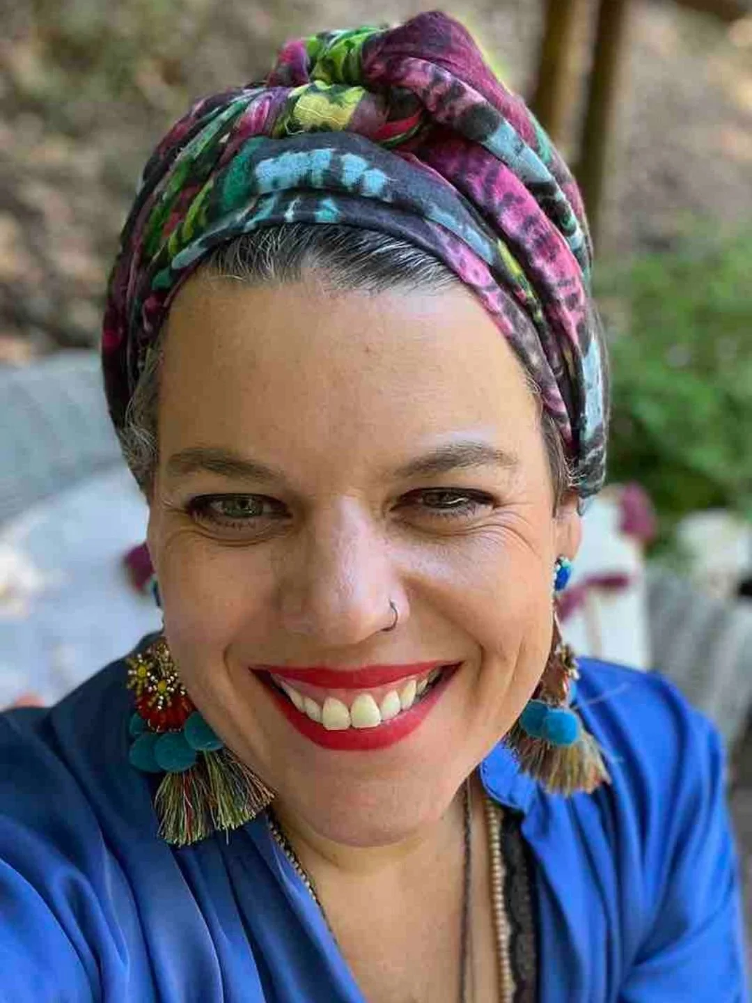 Mulher sorrindo com cabeça envolta em lenço colorido, usando brincos grandes e roupas azuis, ao ar livre.