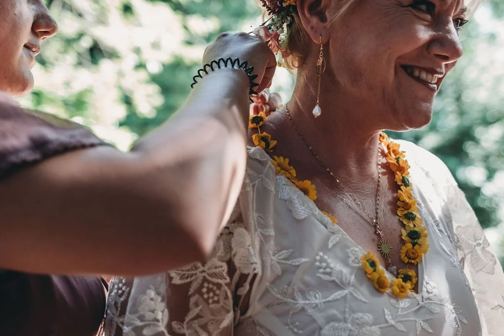 Mulher idosa sorrindo com colar de girassol, usando roupa branca com detalhes em renda, recebendo um colar de flores, possivelmente em uma cerimônia ao ar livre.