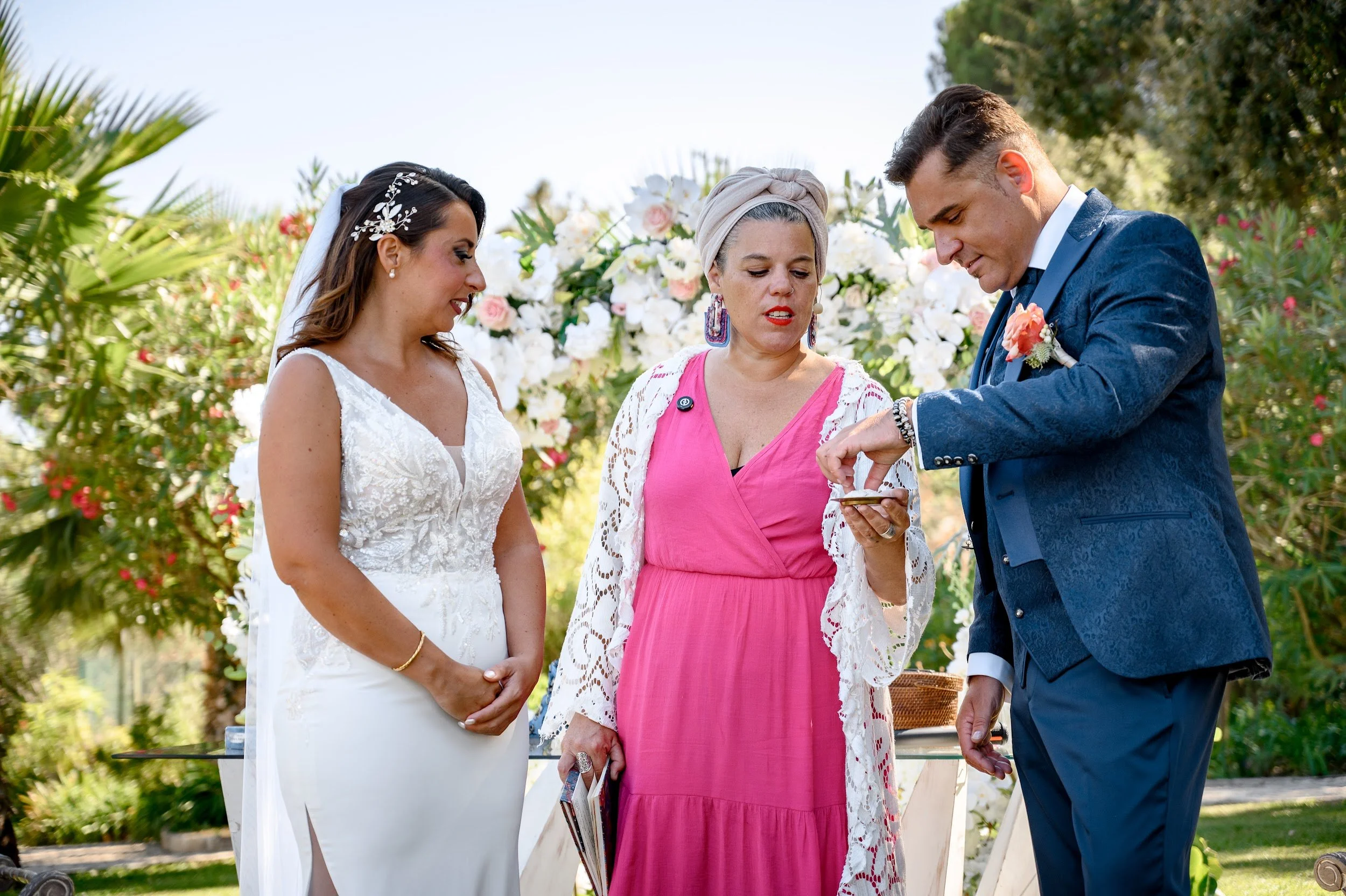 Casal de noivos participando de cerimônia de casamento ao ar livre, com a presença de uma mulher de cabelo cinza e vestido rosa que parece estar realizando o momento.