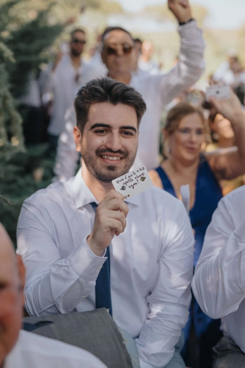 Homem sorridente segurando um bilhete durante um evento ao ar livre, ao fundo outras pessoas, algumas tirando fotos e conversando.
