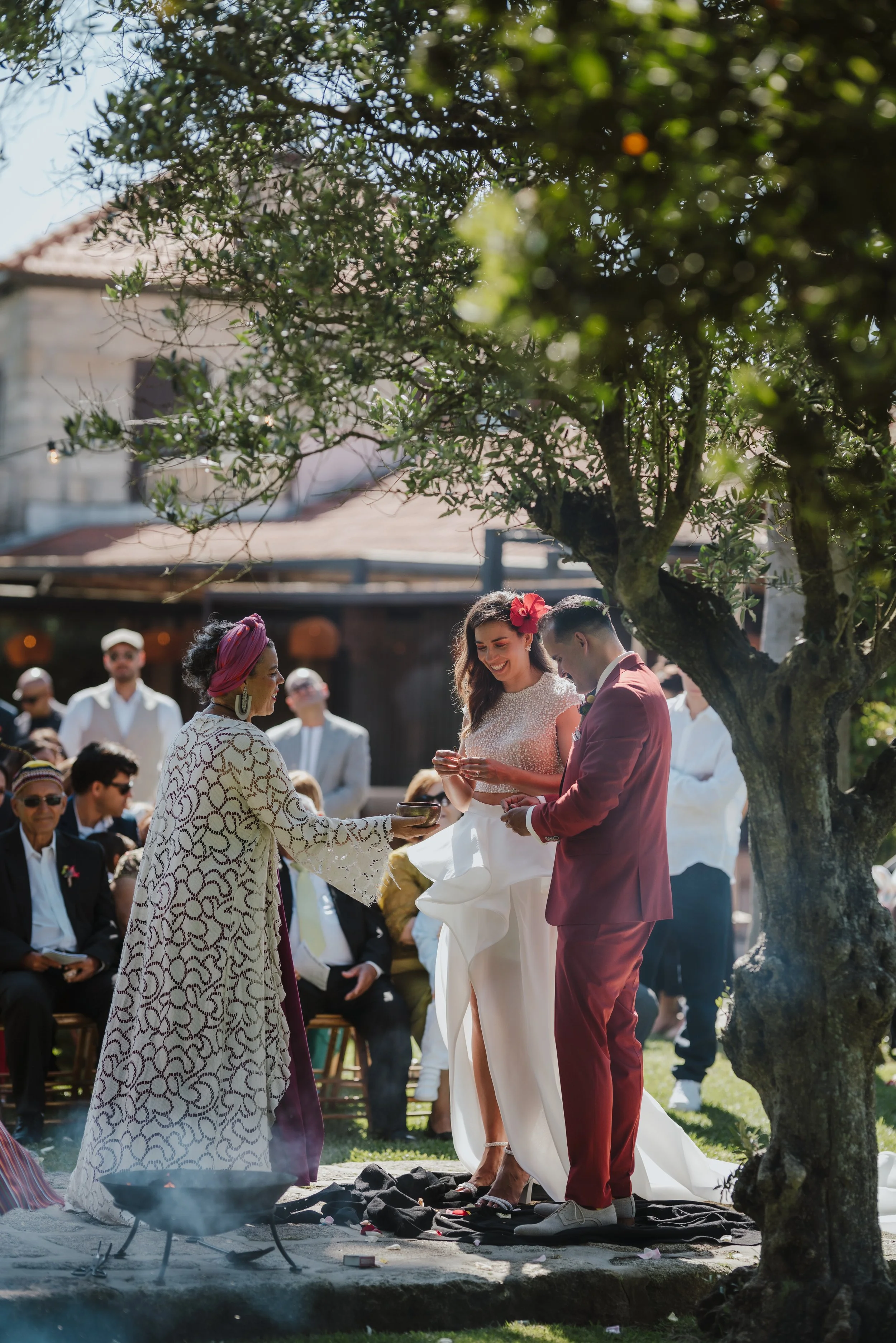 Casamento ao ar livre com convidados assistindo a cerimônia. Uma mulher com vestido branco e flores na cabeça ri enquanto troca votos com um homem de terno rosa. Uma oficiante segura um copo, enquanto uma mulher com turbante e vestido estampado observa, todos sob uma árvore.