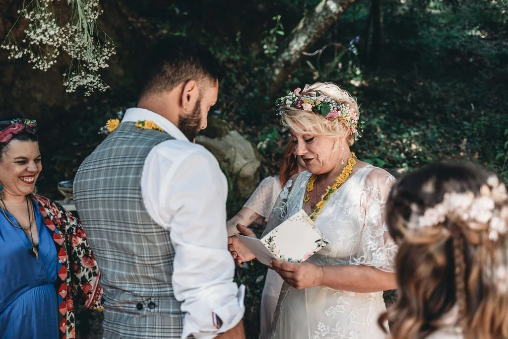 Casamento ao ar livre, cerimônia com uma mulher idosa entregando um papel ao noivo, ao lado de uma mulher sorridente com cabelo trançado e uma mulher com cabelo vermelho, rodeados por árvores e plantas.