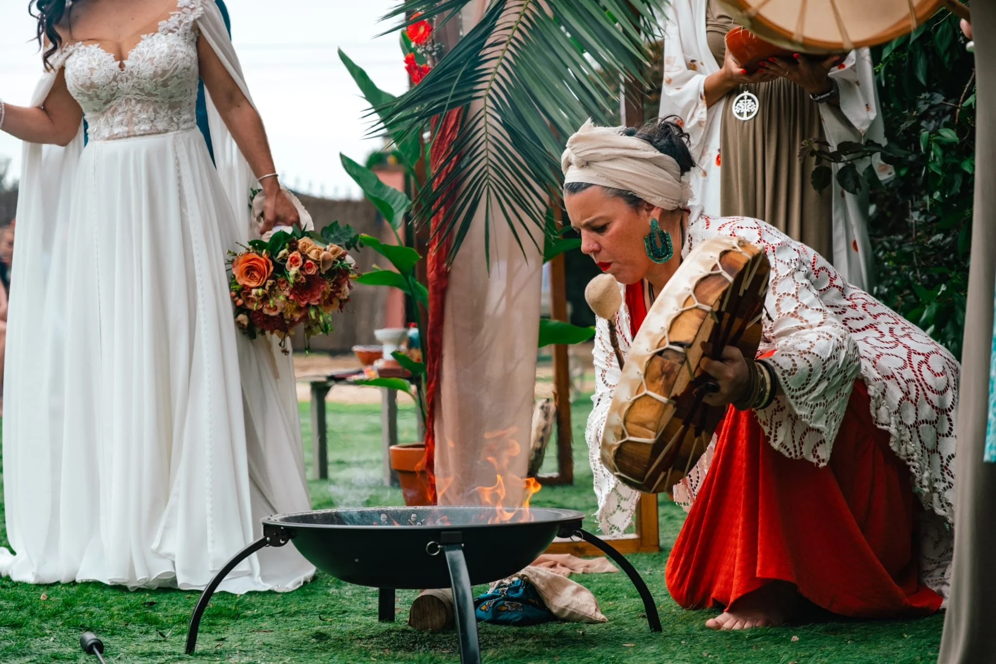 Mulher com vestido de casamento branco ao lado de uma pessoa com roupa tradicional indígena, tocando tambor e balançando um objeto, com fogo em um recipiente