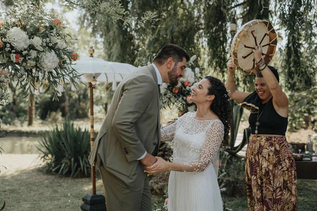 Casal em casamento ao ar livre, com mulher carregando um chapéu de sombrero, segurando as mãos do noivo que está vestido de terno cinza, enquanto a cerimônia é decorada com flores e um guarda-sol branco ao fundo.