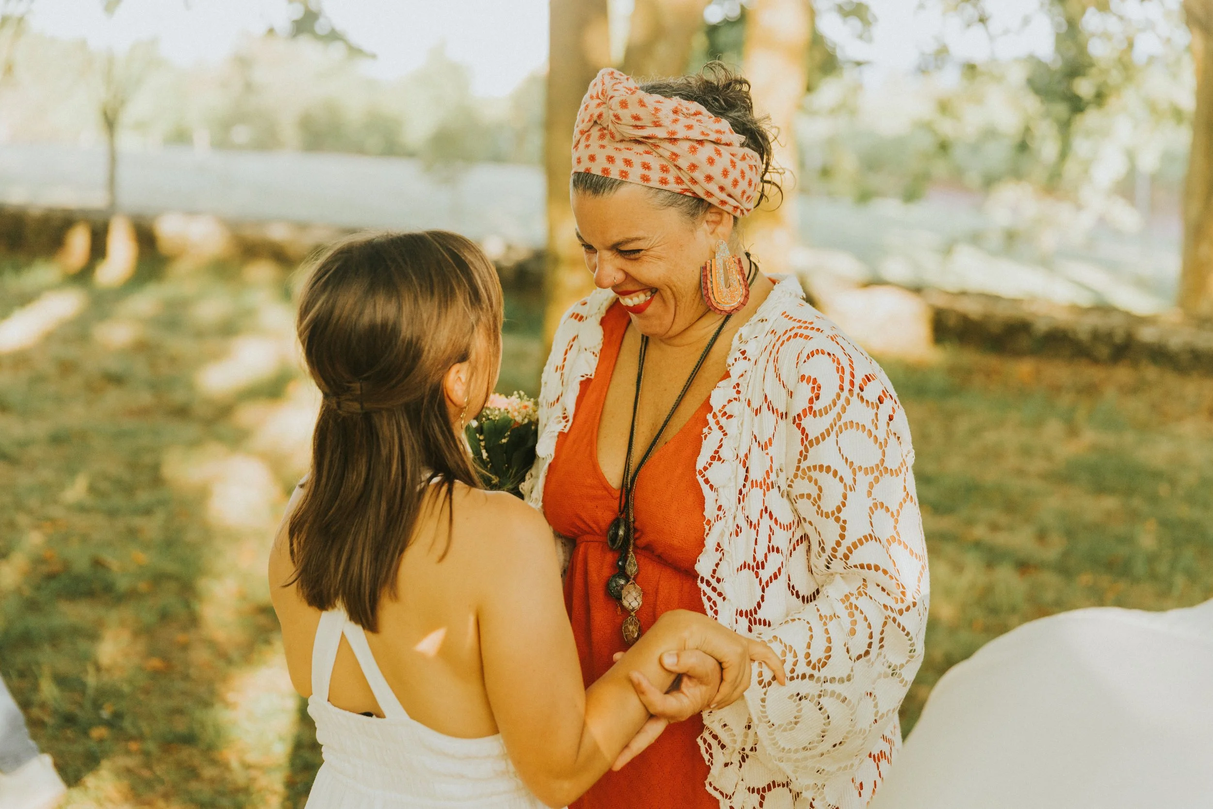 Mulher sorridente com cabeça envolta em lenço floral, usando brincos grandes, interagindo afetuosamente com criança de cabelos longos e cabelo preso, em ambiente ao ar livre com árvores no fundo.