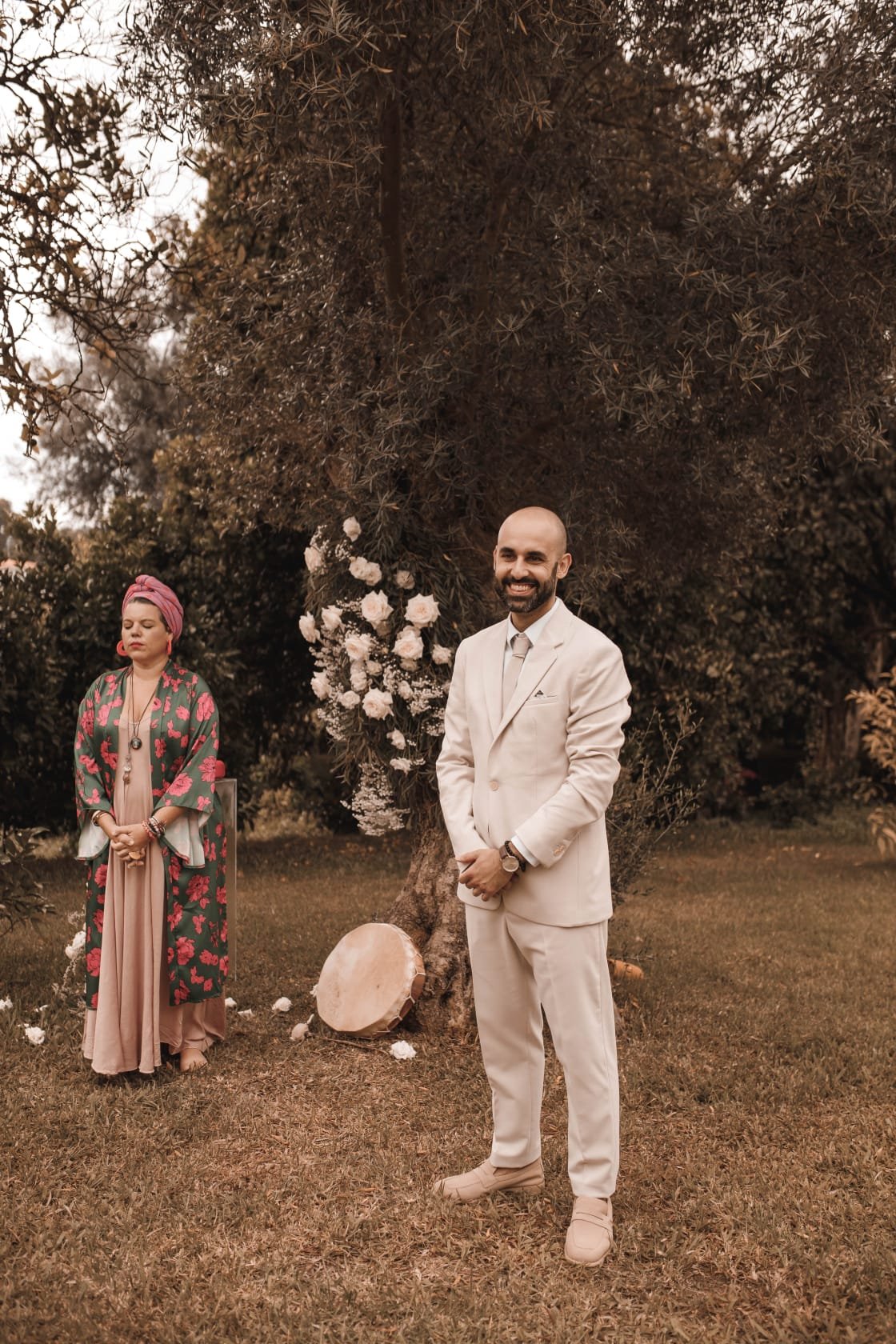 Homem sorrindo vestido de terno branco, mulher com vestido longo e roupão floral, em um ambiente ao ar livre com árvores e flores.