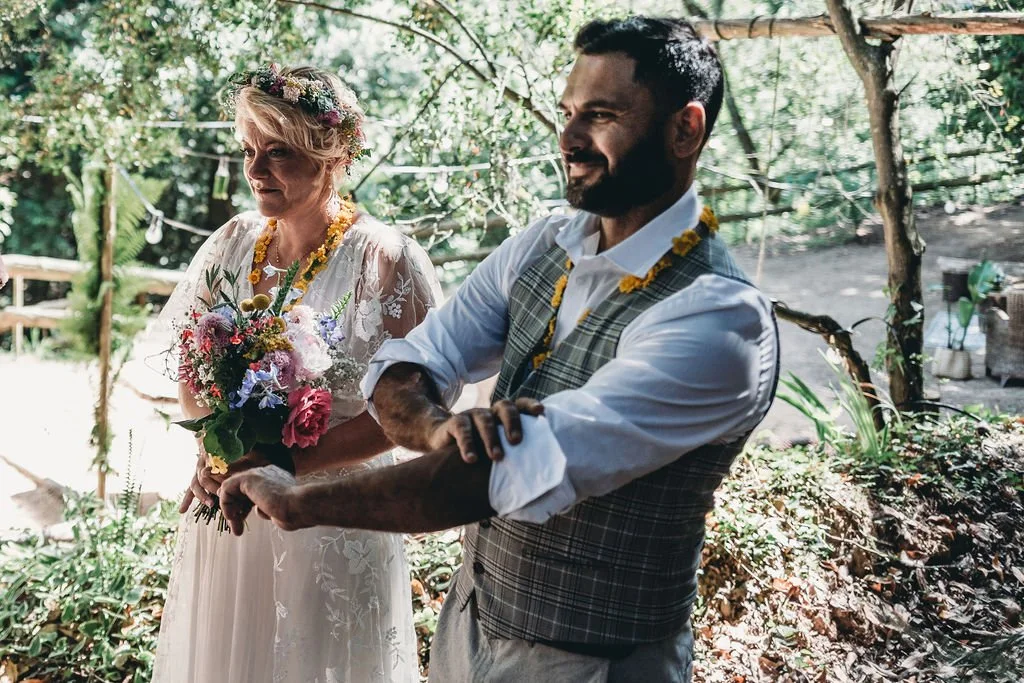 Casal de casamento no jardim, com a mulher segurando um buquê de flores coloridas e o homem com braços cruzados, ambos usando colares de flores.