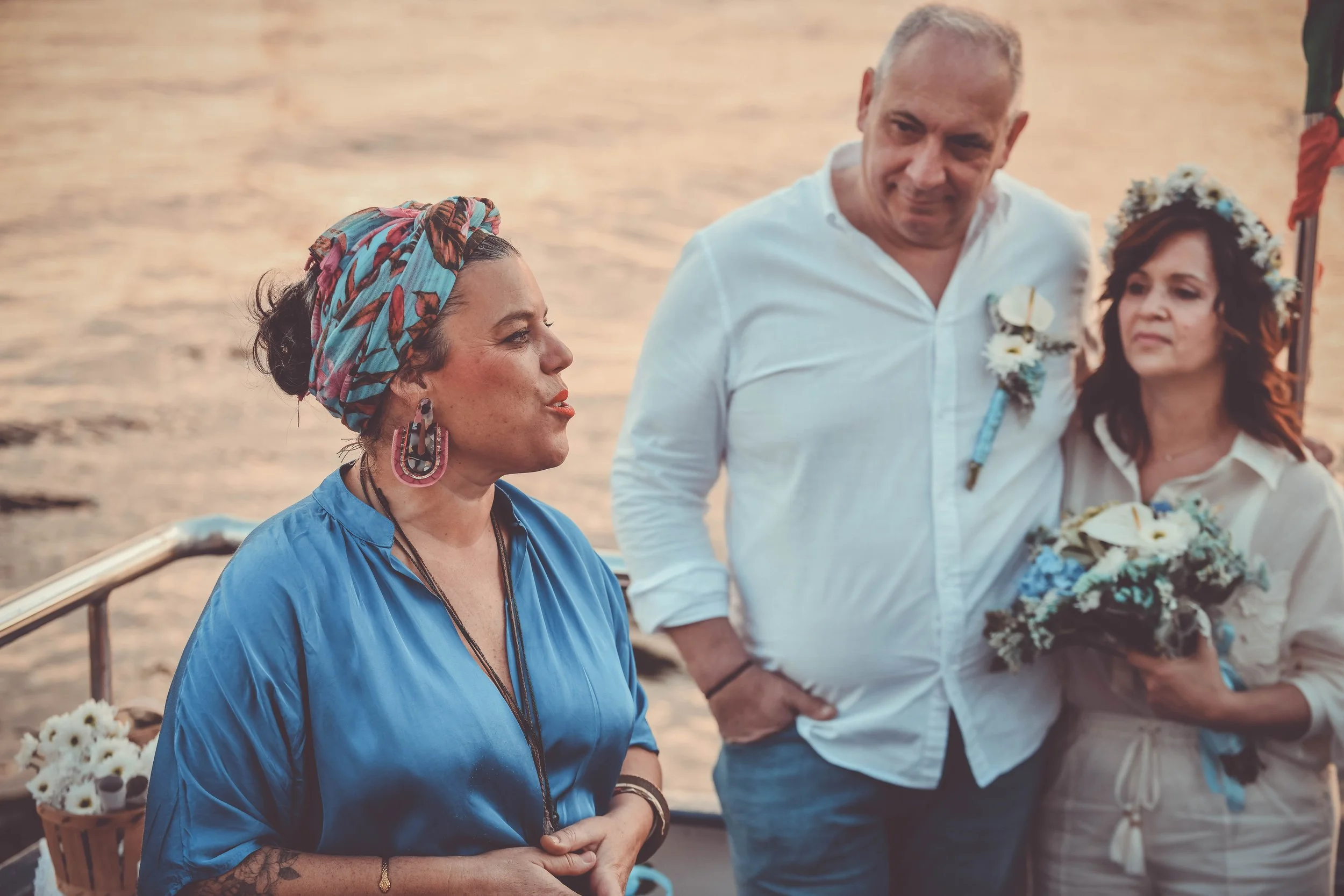 Mulher sorridente com um turbante colorido e brincos grandes, ao lado de um homem sorridente com um lenço branco, em uma cerimônia ao ar livre com uma mulher segurando um buquê de flores.