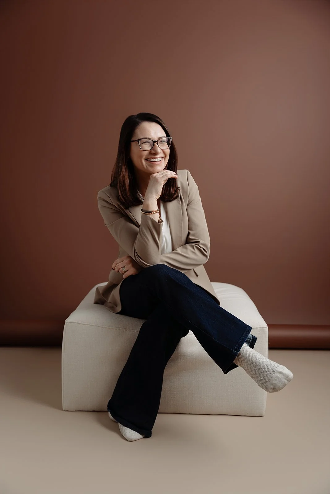 A woman with glasses, wearing a beige blazer, white shirt, and black pants, sitting on a beige ottoman against a brown background, smiling and laughing.