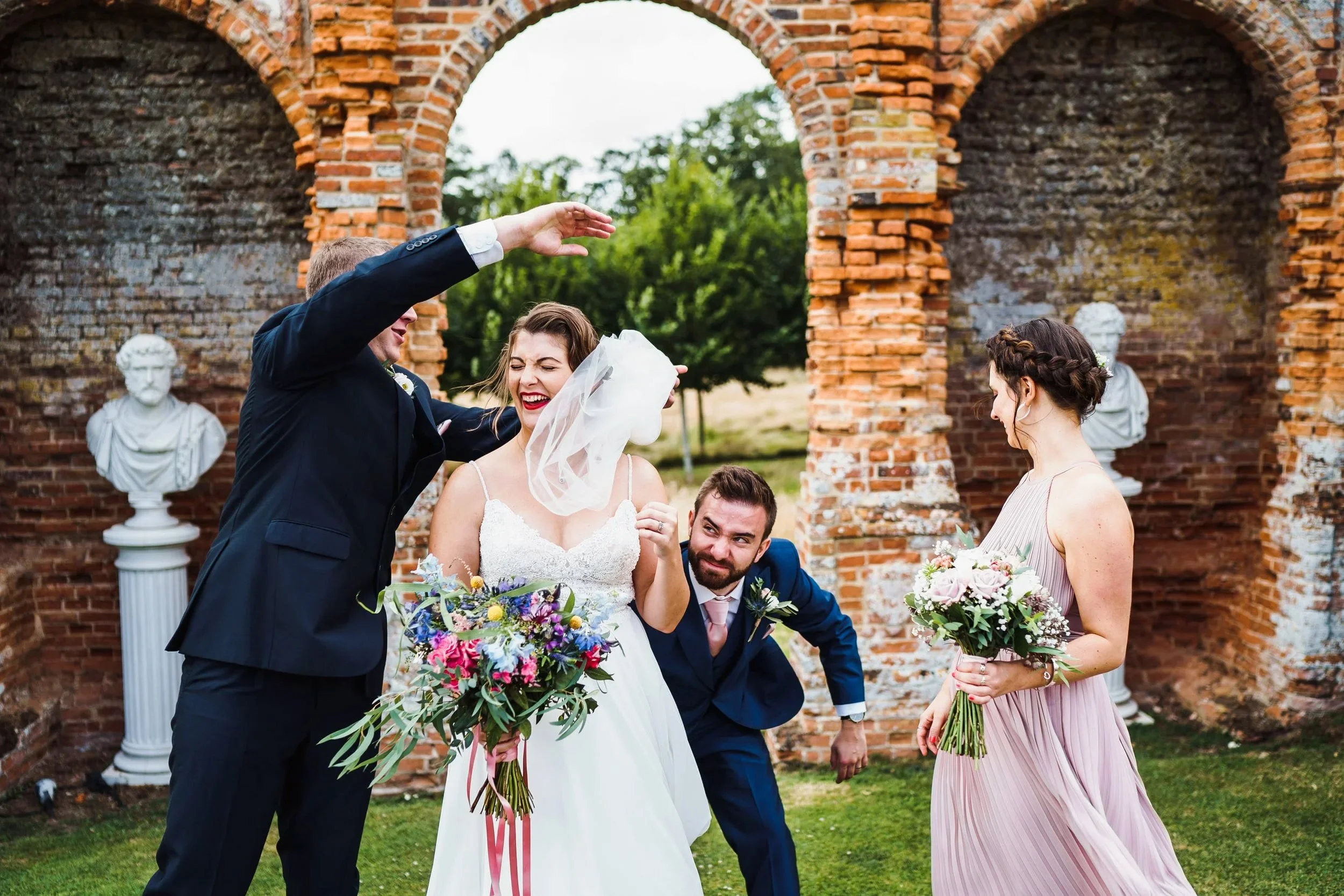 A windy day during the wedding group photos at Oxnead Hall sees the groom and best man wrangle with the brides dress and veil. The bride is laughing as her chief bridesmaid watches on.