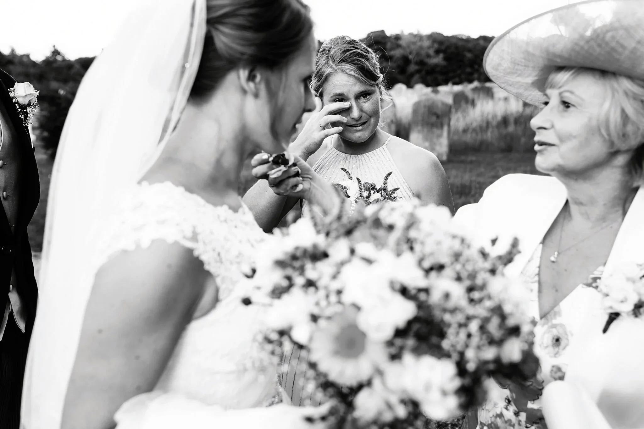 A bridesmaid, in the centre of the picture, framed either side by the bride and her mother,  wipes a tear from her eye as she is overcome with emotion.