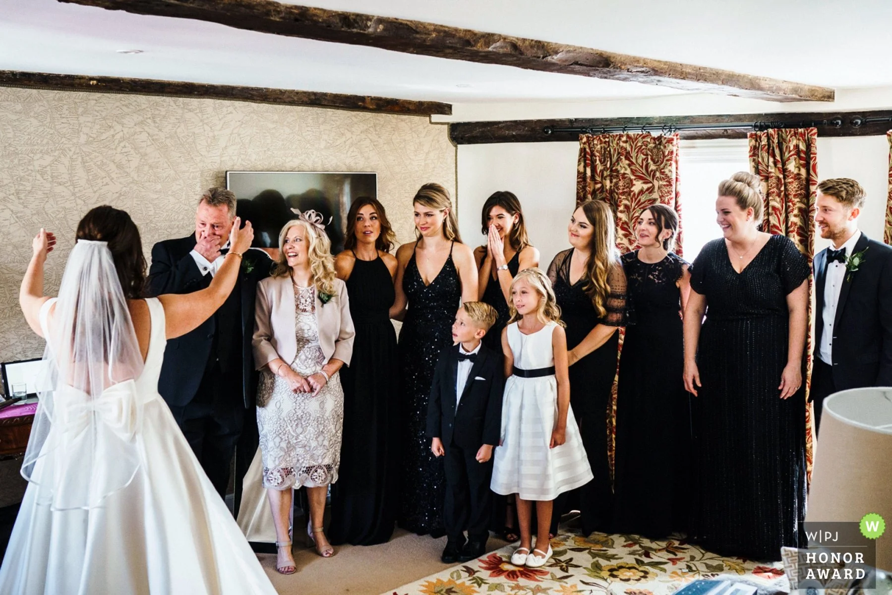 A bride with arms open wide toward her father as he wipes away tears upon seeing her. Other relatives and guests look on with tears and smiles on their faces also.
