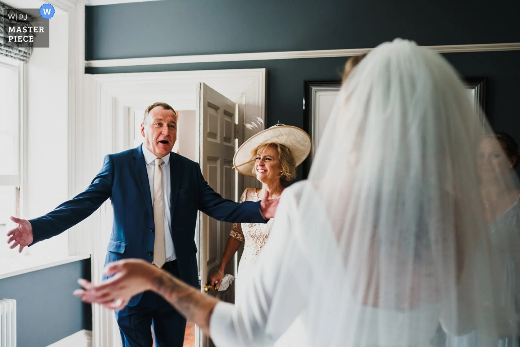 Father of the bride opens his arms wide to embrace his daughter when he sees her for the first time before walking her down the aisle. The brides mother watches on with a beaming smile. 