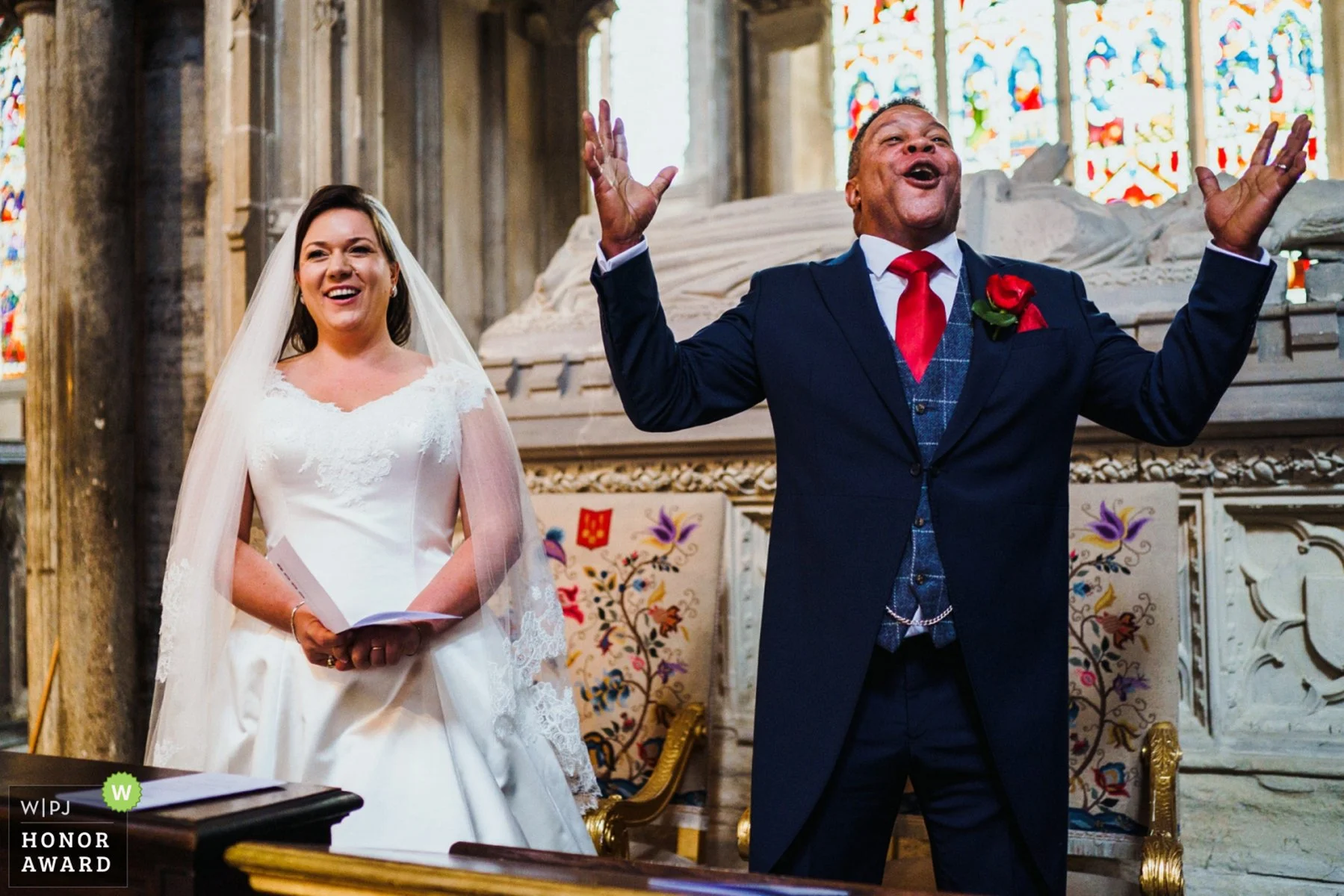 A groom holds his arms out wide and has a huge smile on his face as he sings hymns at Ely Cathedral. His newlywed wife smiles and sings alongside. 