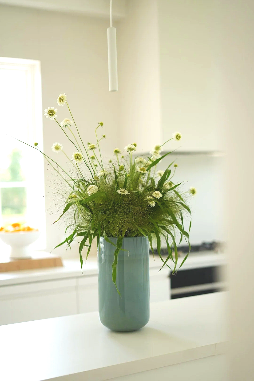 A tall blue vase with a bouquet of white wildflowers and green foliage on a white surface in a bright, modern kitchen.