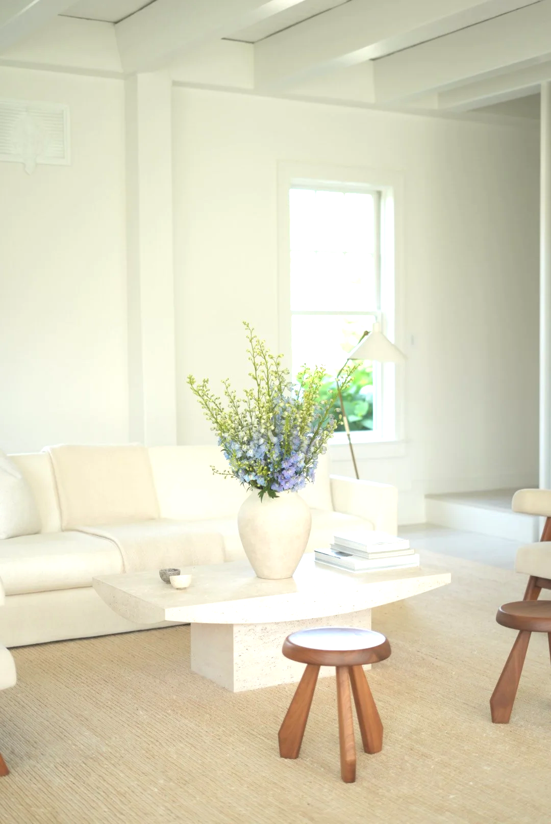 Bright, minimalist living room with a white sofa, a white coffee table with a large floral arrangement, a small stool, a floor lamp, and a window with sunlight streaming in.