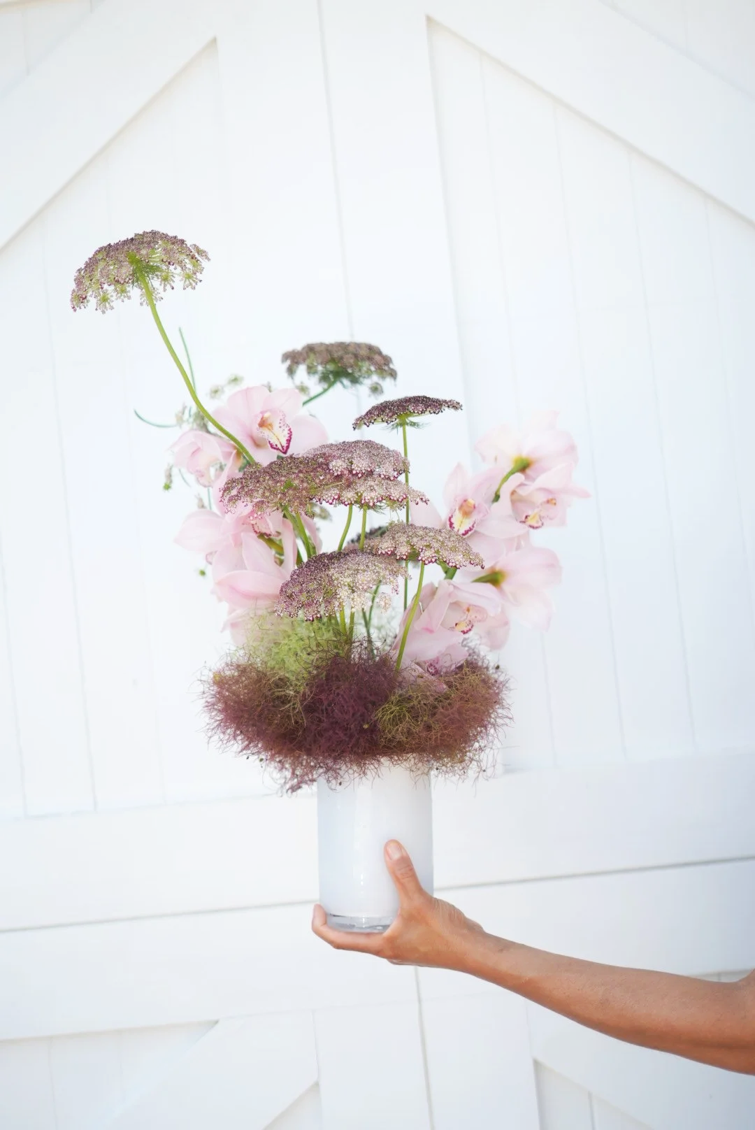 Hand holding a white vase with a floral arrangement of pink orchids, purple-tinted flowers, and decorative moss against a white wooden background.