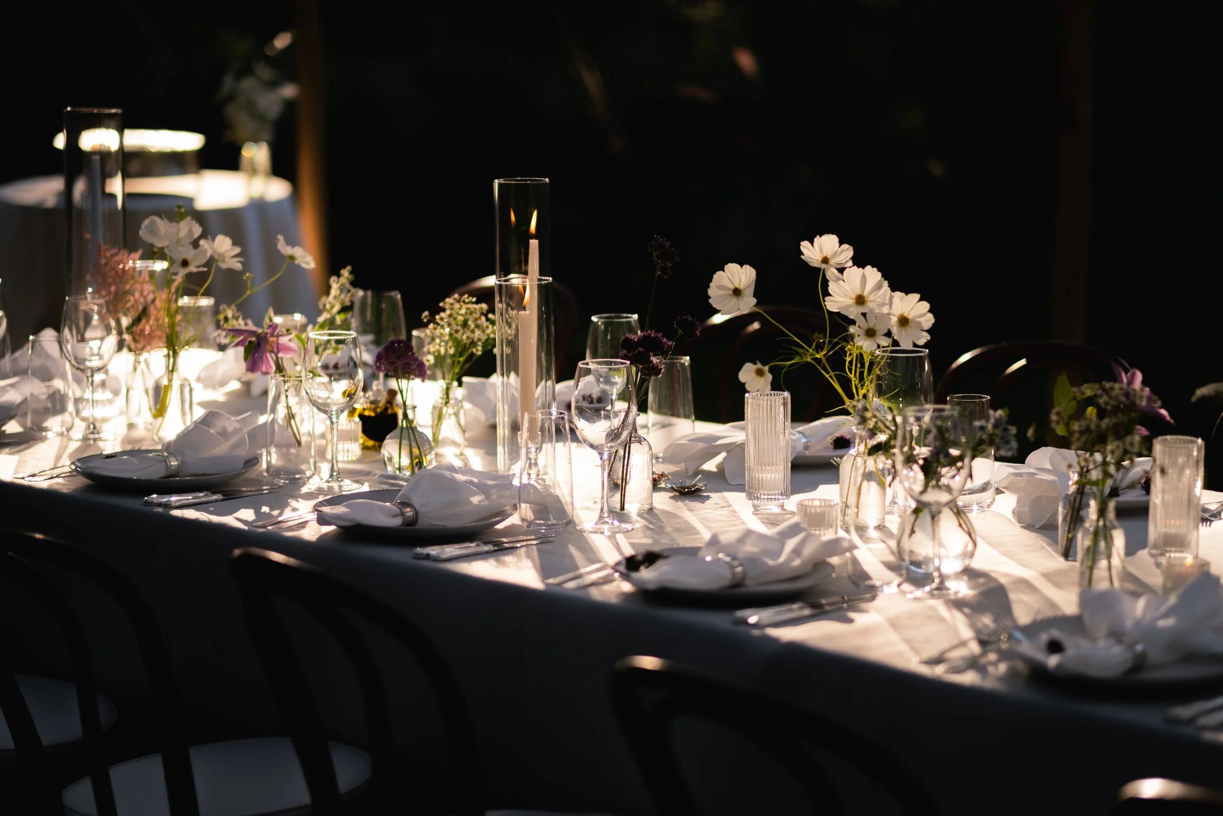 At LongHouse Reserve in East Hampton, floral centerpieces in vases, glassware, and napkins set for a meal, illuminated by candles and sunlight. Floral, flower wedding and event design based in the North Fork and Hamptons.