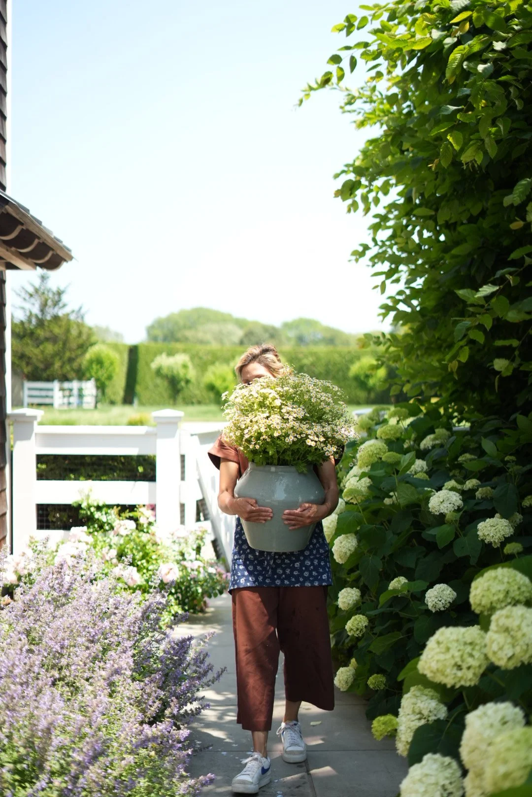 A woman carrying a large white pot of white and yellow flowers while walking along a garden path lined with flowering plants and green shrubbery.