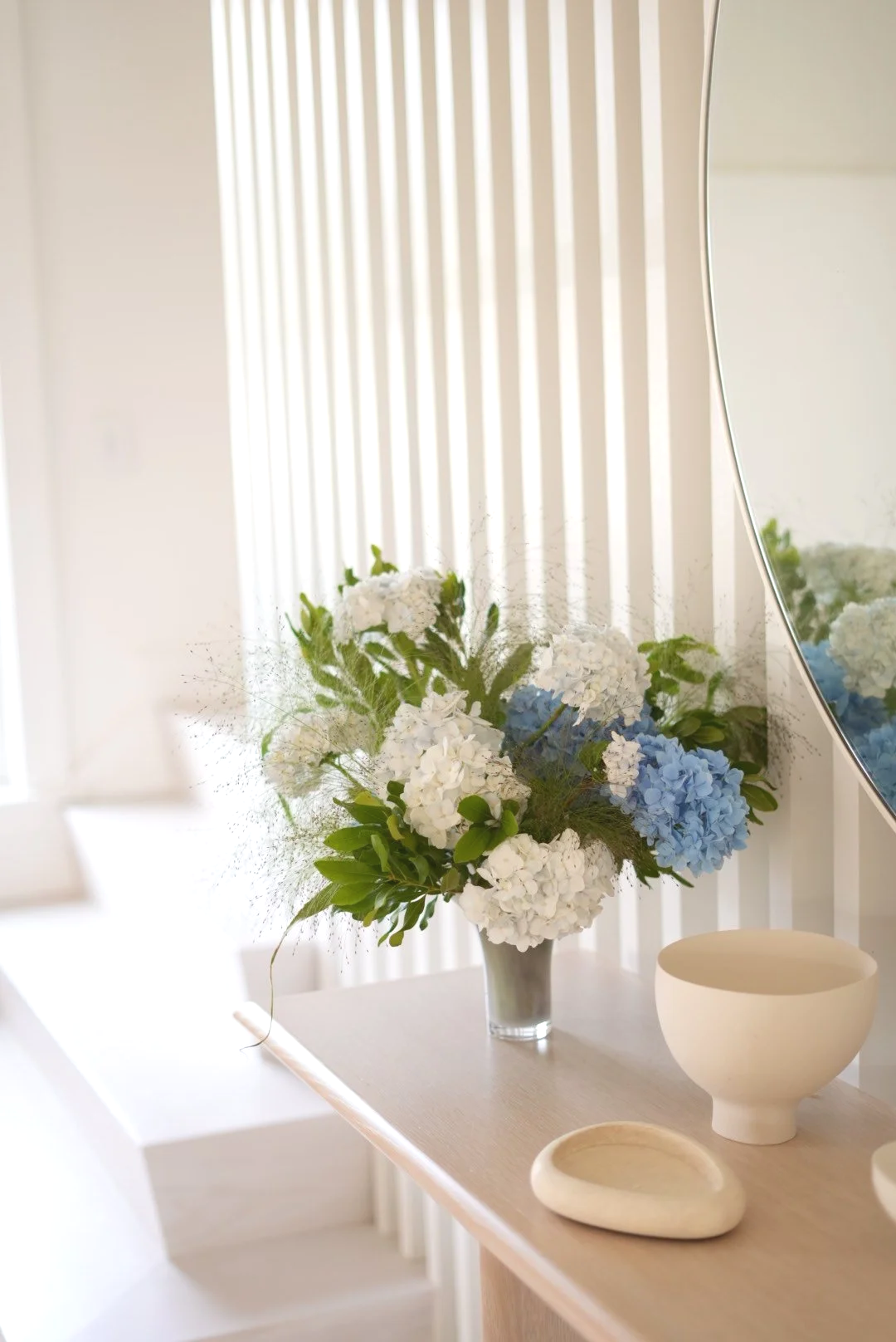 A vase with white and blue hydrangeas on a beige table, with decorative white bowls nearby. A mirror reflects part of the flower arrangement and light filtering through vertical blinds.