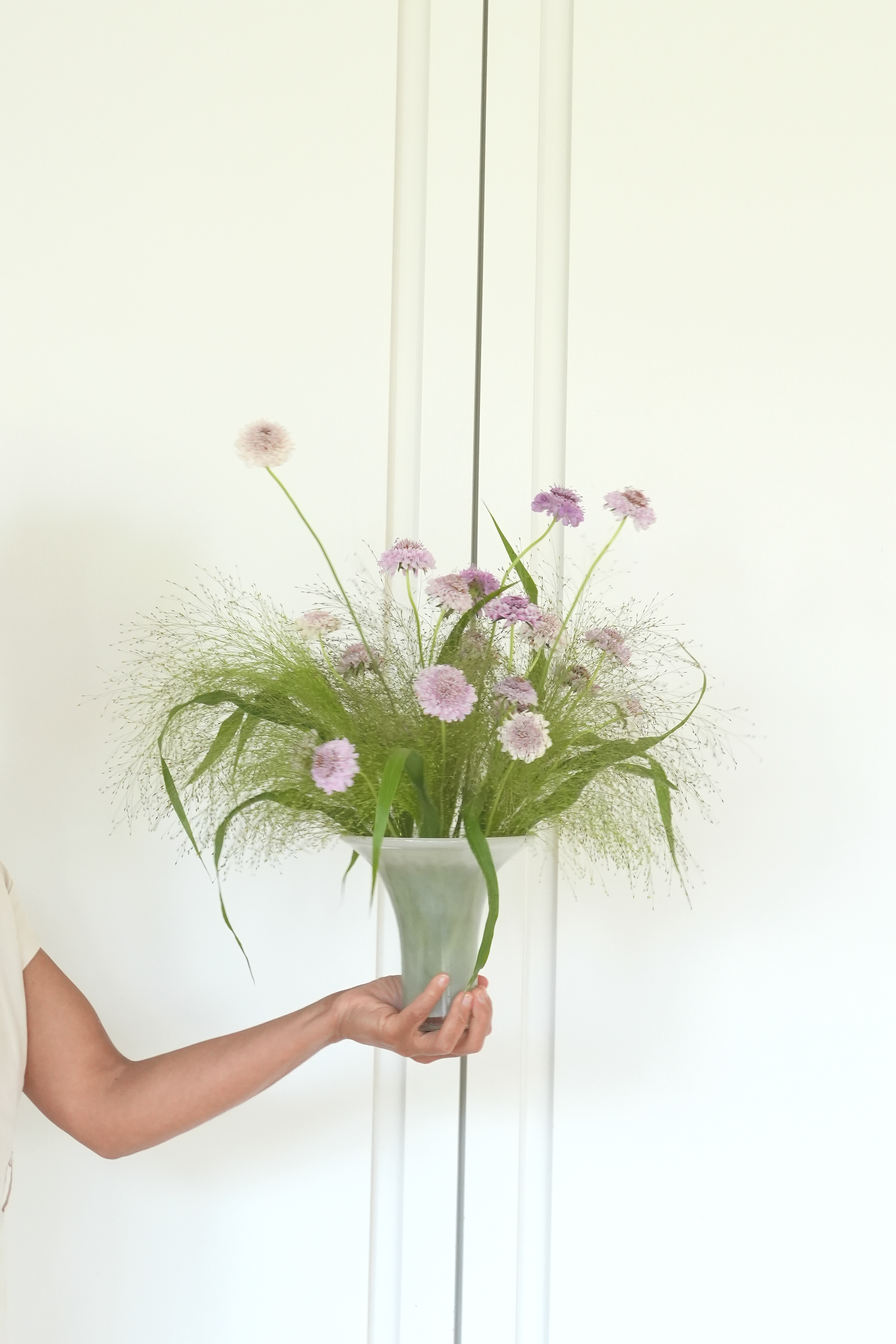 A person is holding a tall, white vase with a flower arrangement of pink and purple flowers and green foliage against a plain, white background.