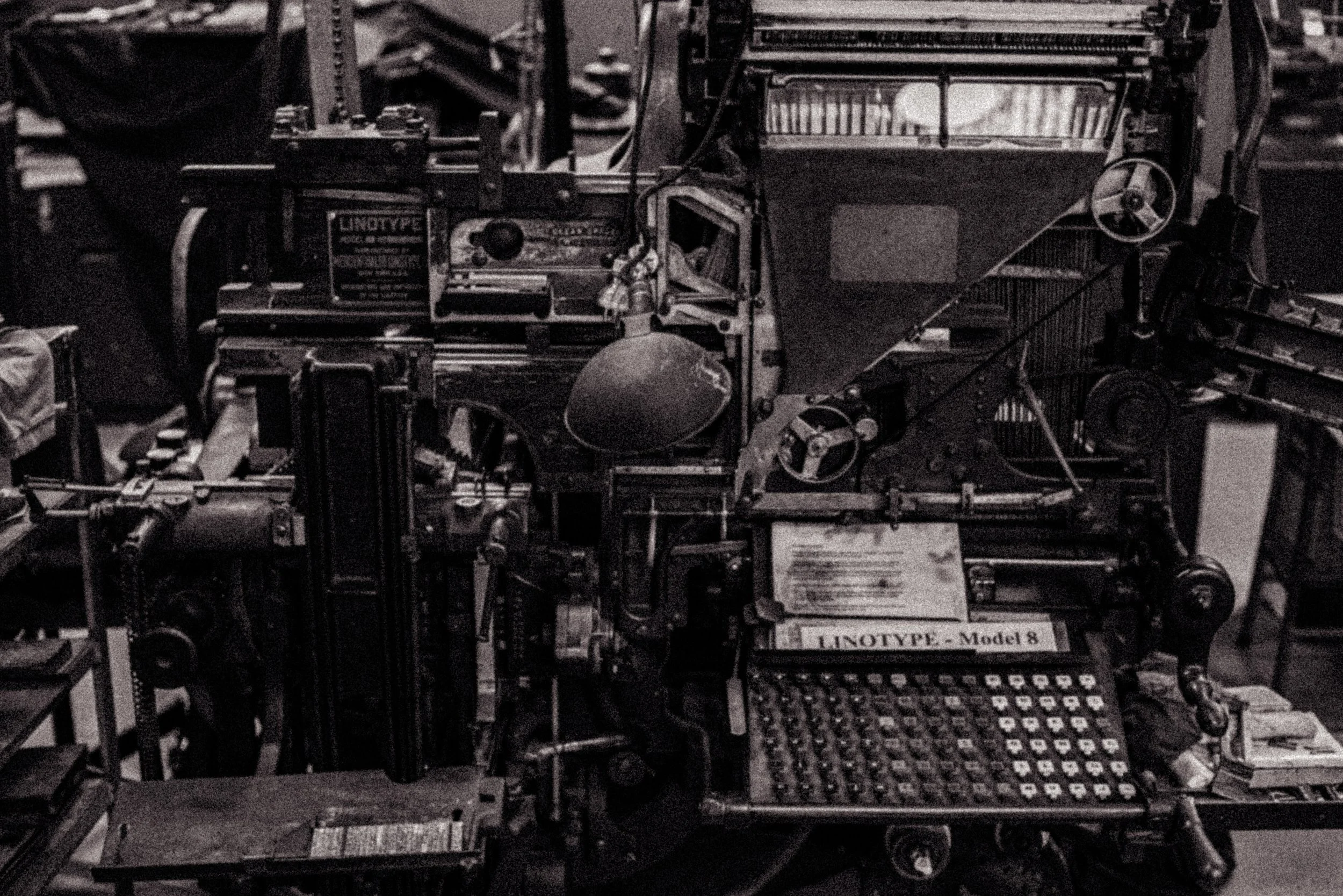A black and white photo of a vintage mechanical device labeled 'LINOTYPE - Model 8', with various gears, levers, and metal parts, possibly used for printing or typesetting.