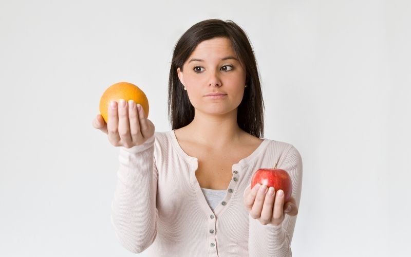 woman weighing an orange in her right hand and an apple in her left hand, looking at the orange.