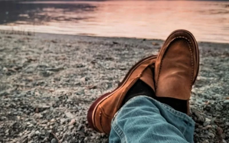 Man crossing feet with brown slipons and jeanson pebble strewn beach