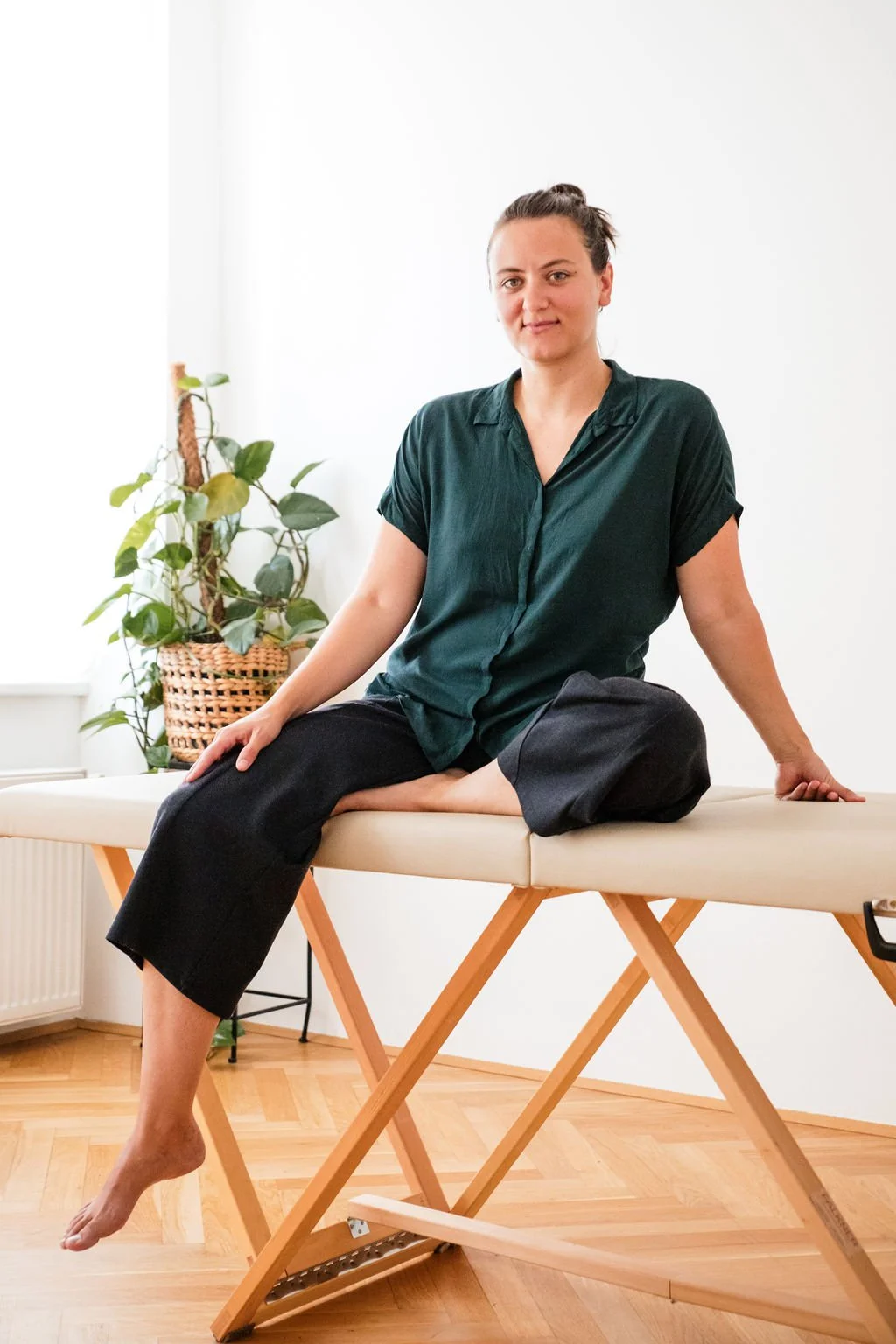 Körpertherapeutin sitzt auf einer Massageliege in ihrem hellen Praxisraum mit Holzfußboden und Pflanze im Hintergrund.