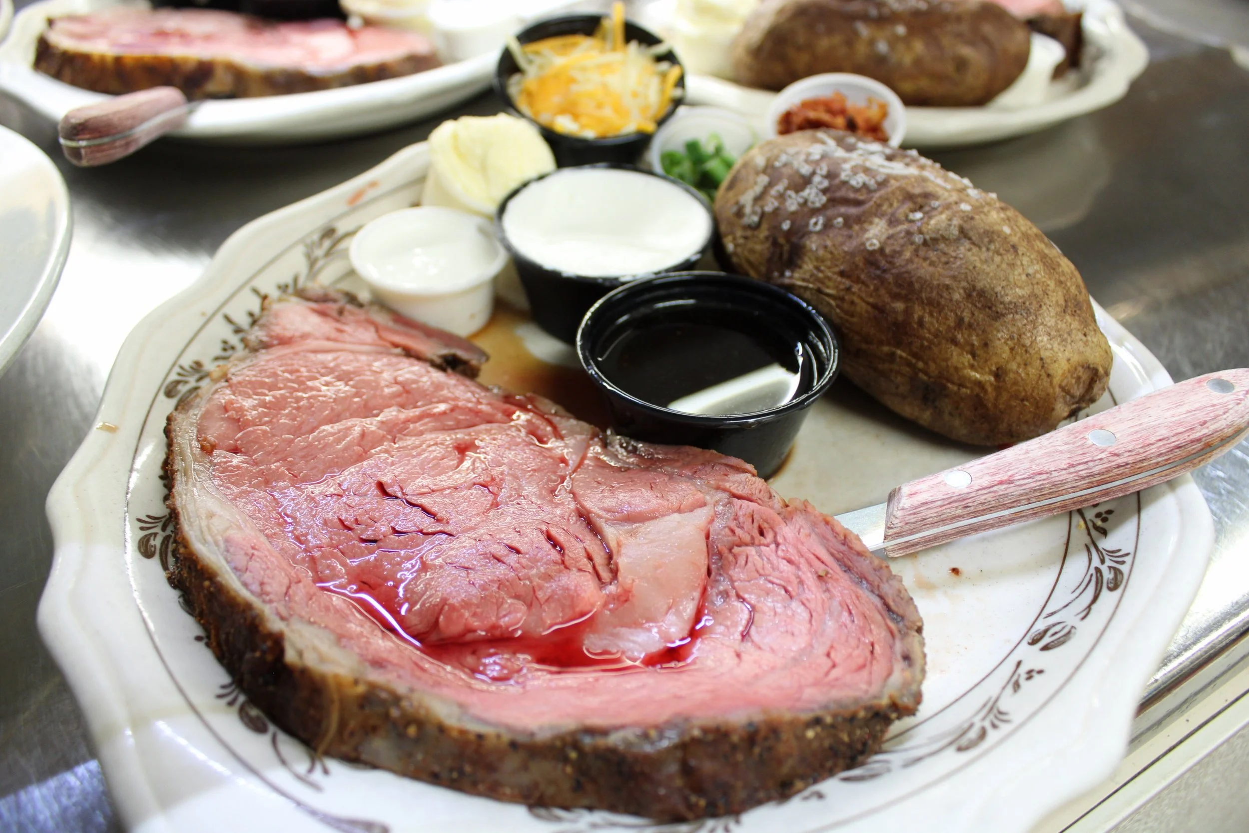 A photo of the 8-ounce prime rib dinner at the Sunset Cafe and Loose Wolf Saloon. The dinner is plated with a baked potato and small cups of au jus, sour cream, creamed horseradish, cheese, chives, and bacon.