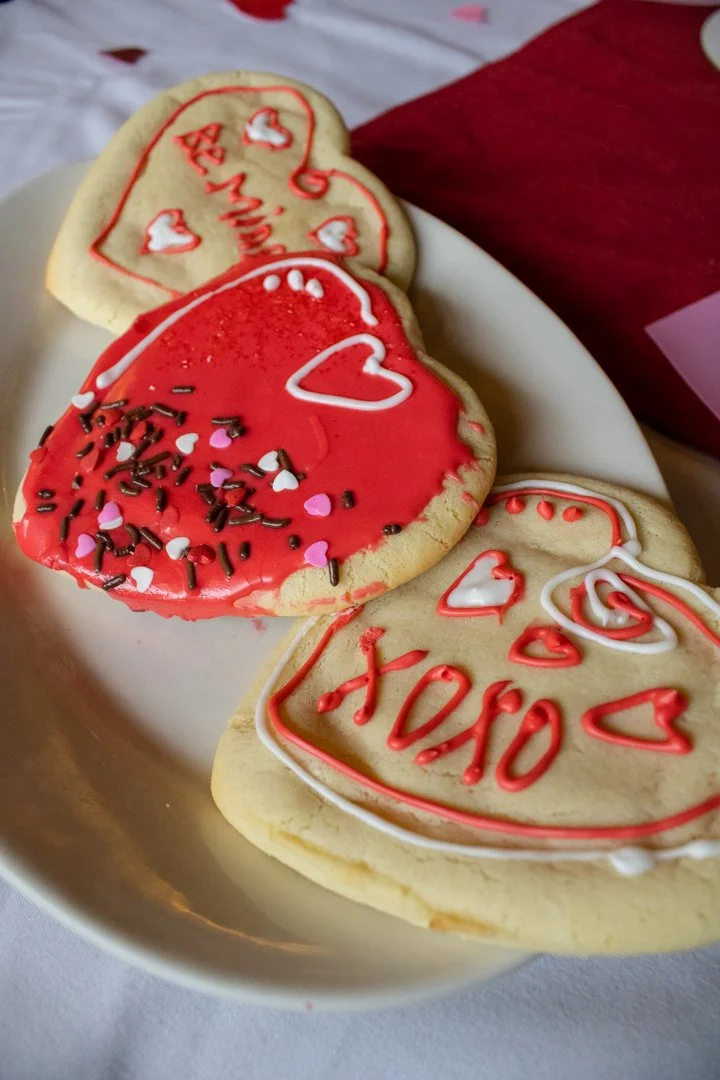 Photo of three heart-shaped sugar cookies on a plate at the Sunset Cafe and Loose Wolf Saloon. The middle cookie has red icing and sprinkles. The left cookie has "Be Mine" written in icing, and the right cookie has "xoxo" written in icicing.