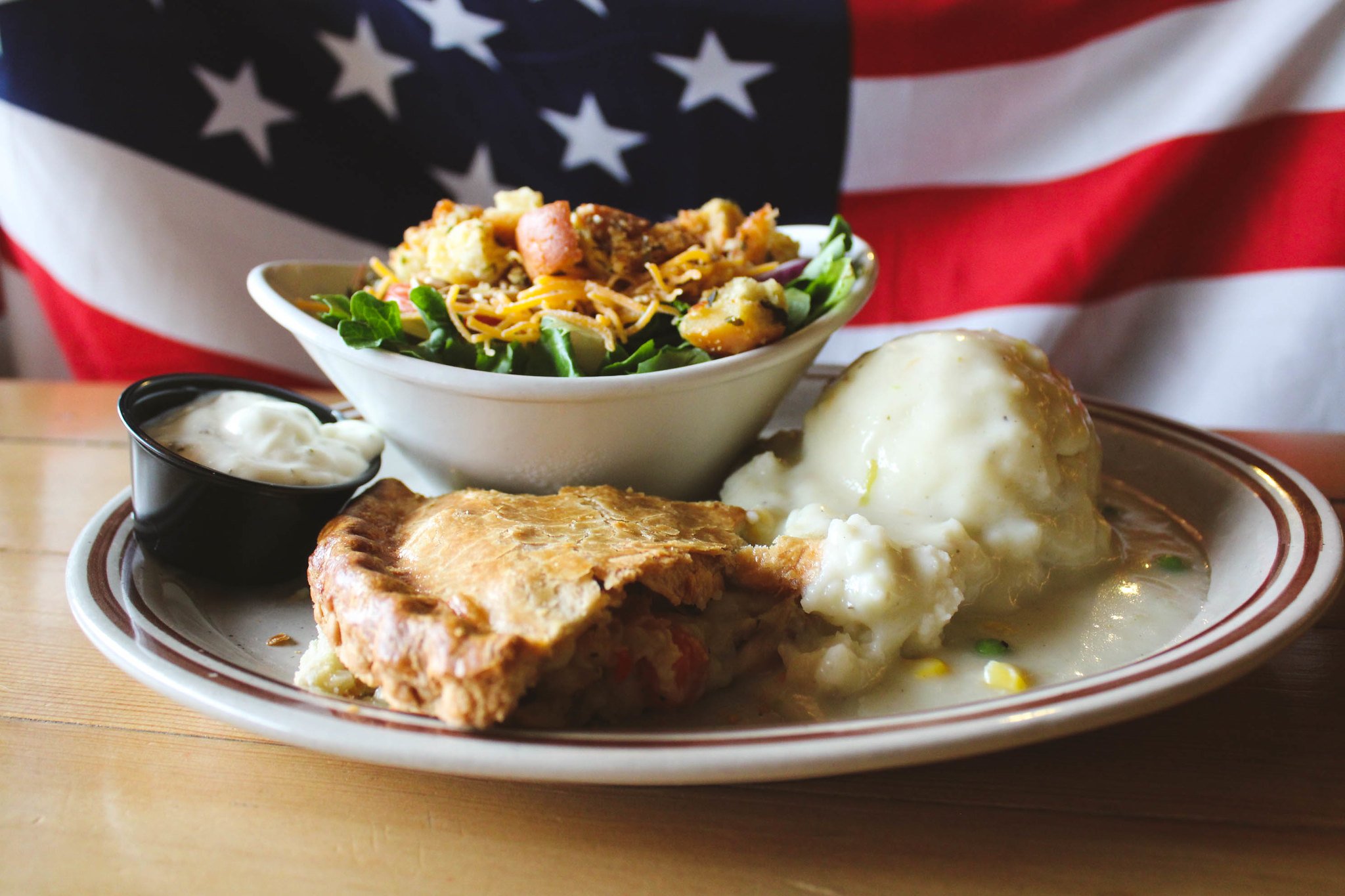 A photo of a slice of chicken pot pie at the Sunset Cafe and Loose Wolf Saloon. The pot pie is plated with mashed potatoes and gravy and a green salad. The plate is on a wooden table, and there is an American flag in the background.