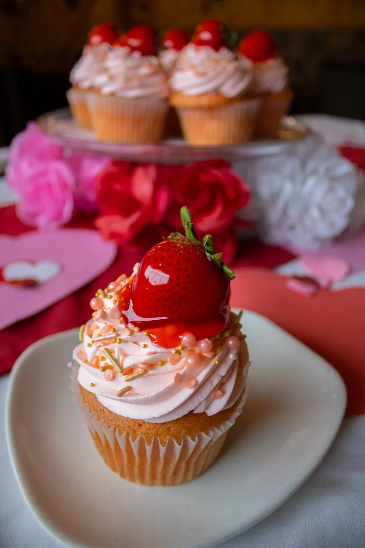 Photo of a strawberry cupcake topped with icing and a fresh strawberry at the Sunset Cafe and Loose Wolf Saloon. The cupcake sits on a plate, and in the background, there are more strawberry cupcakes on an elevated plate. The table has white, pink, a