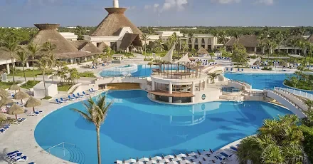 A large outdoor swimming pool with a circular section and a separate lazy river, surrounded by lounge chairs and palm trees, with a resort building and thatched-roof structures in the background.