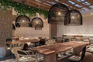 Interior of a restaurant with wooden tables, black chairs, woven pendant lights, and a stone accent wall with a patterned wallpaper.