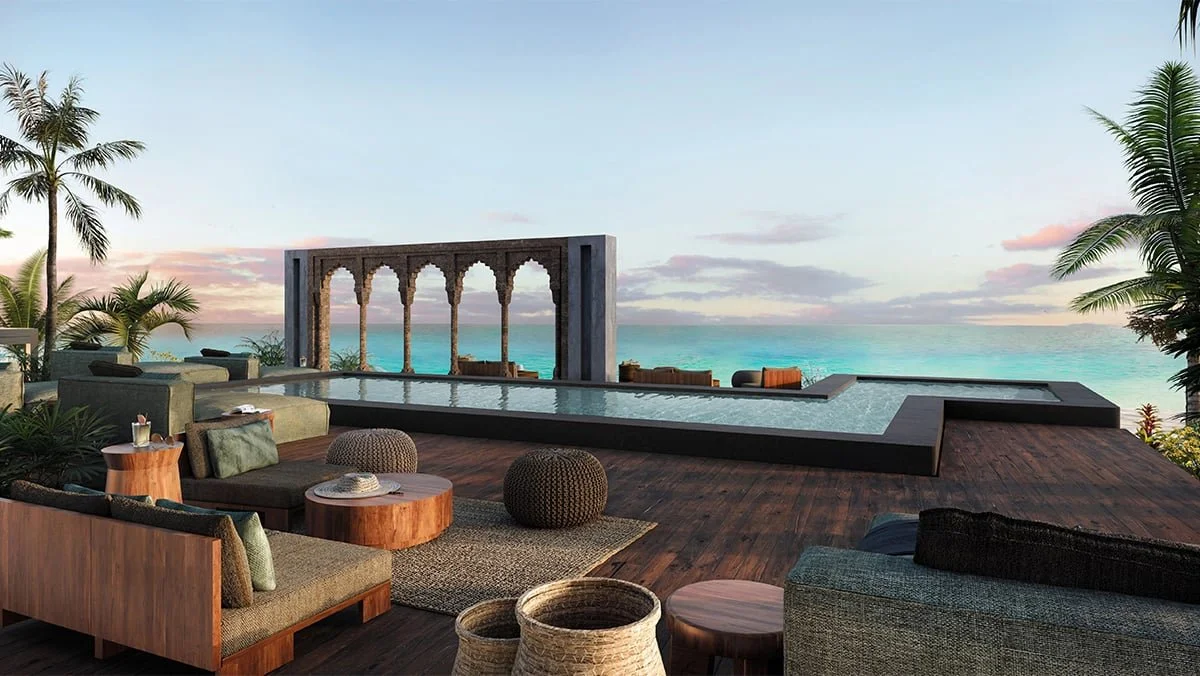 Luxury outdoor lounge area with wooden flooring, seating, and cushions overlooking a infinity pool, palm trees, and ocean at sunset.