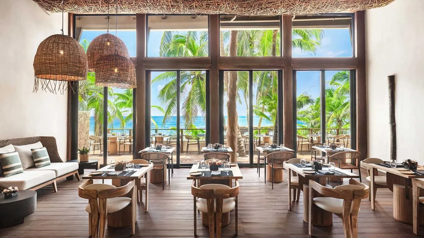 Interior view of a beachside restaurant with large windows showing palm trees and the ocean, wooden tables and chairs, woven pendant lights hanging from the ceiling, and a cozy seating area with cushions.