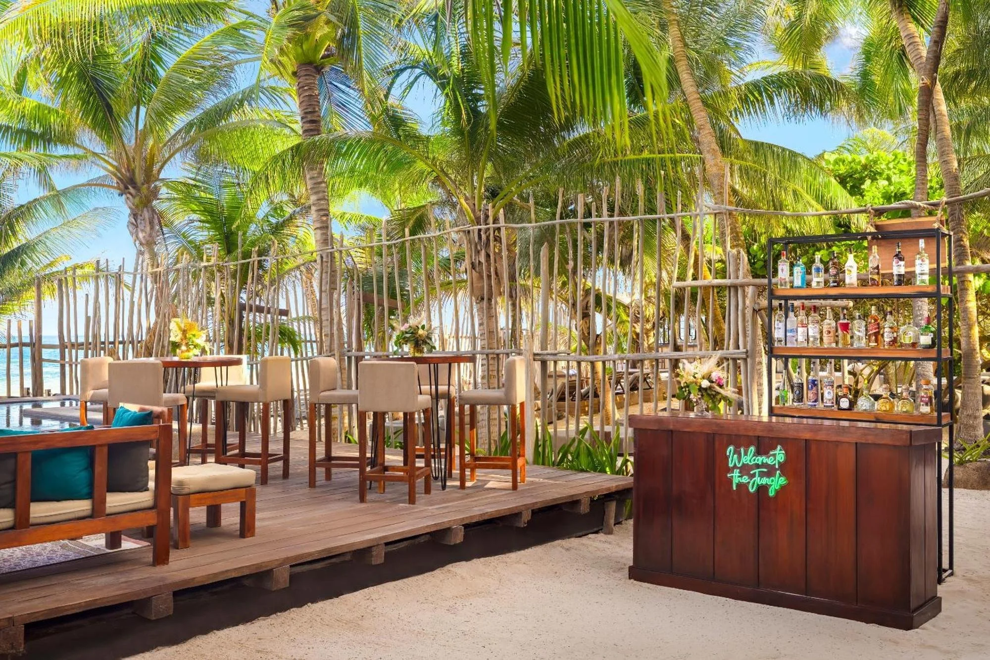 A tropical outdoor bar on a wooden deck surrounded by palm trees, with high chairs and small tables. There is a bar counter with a sign that reads 'Welcome to the Jungle' and a shelf with various bottles of alcohol.