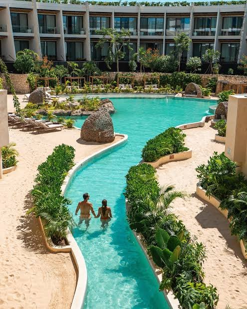 Two people swimming in a winding pool at a hotel or resort, surrounded by greenery and rocks, with a modern building in the background.